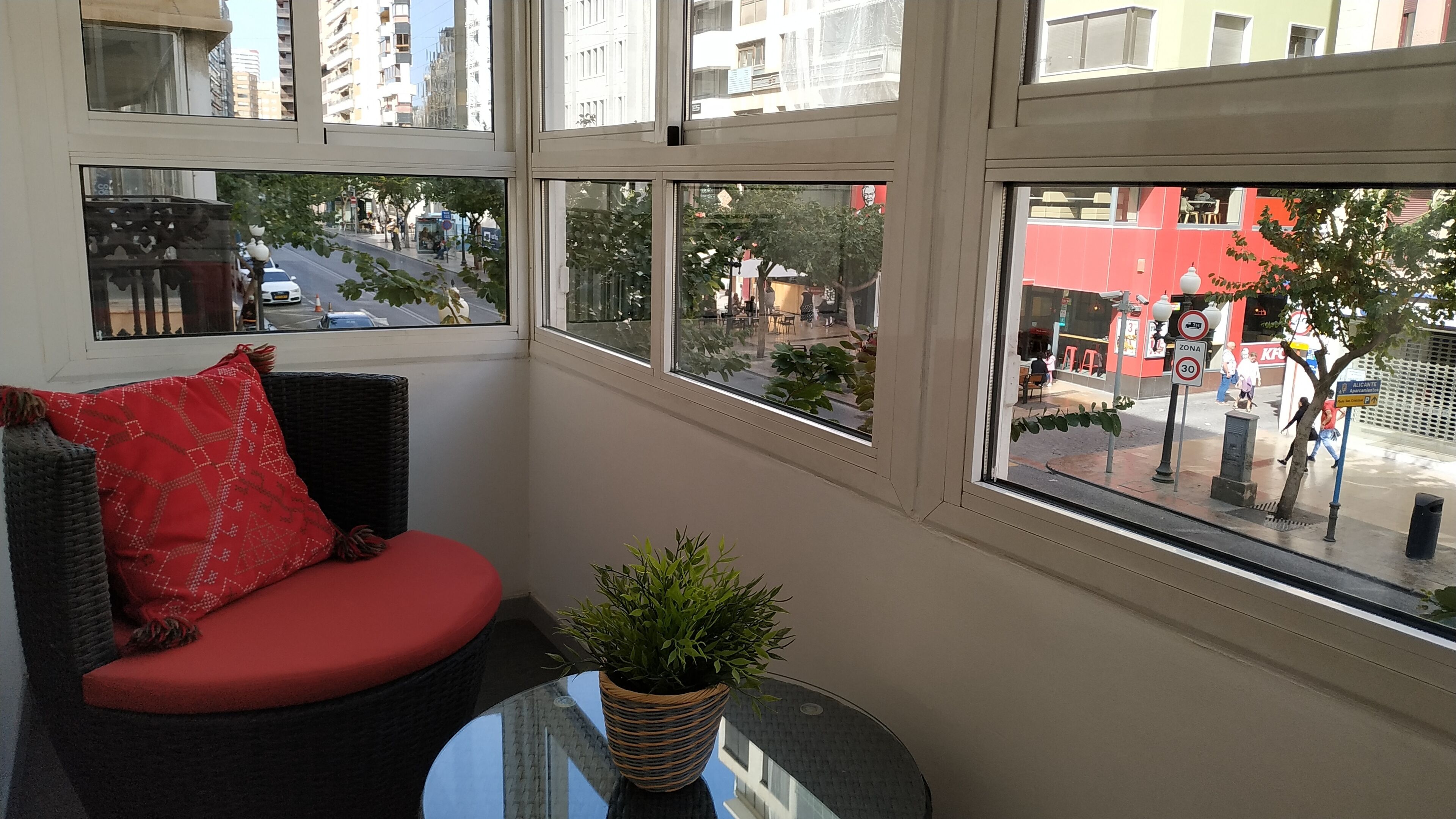 Ein Zimmer mit einem Korbssessel, einem kleinen Tisch mit einem Topfpflanze und großen Fenstern mit Blick auf die Straße und die umliegenden Gebäude.