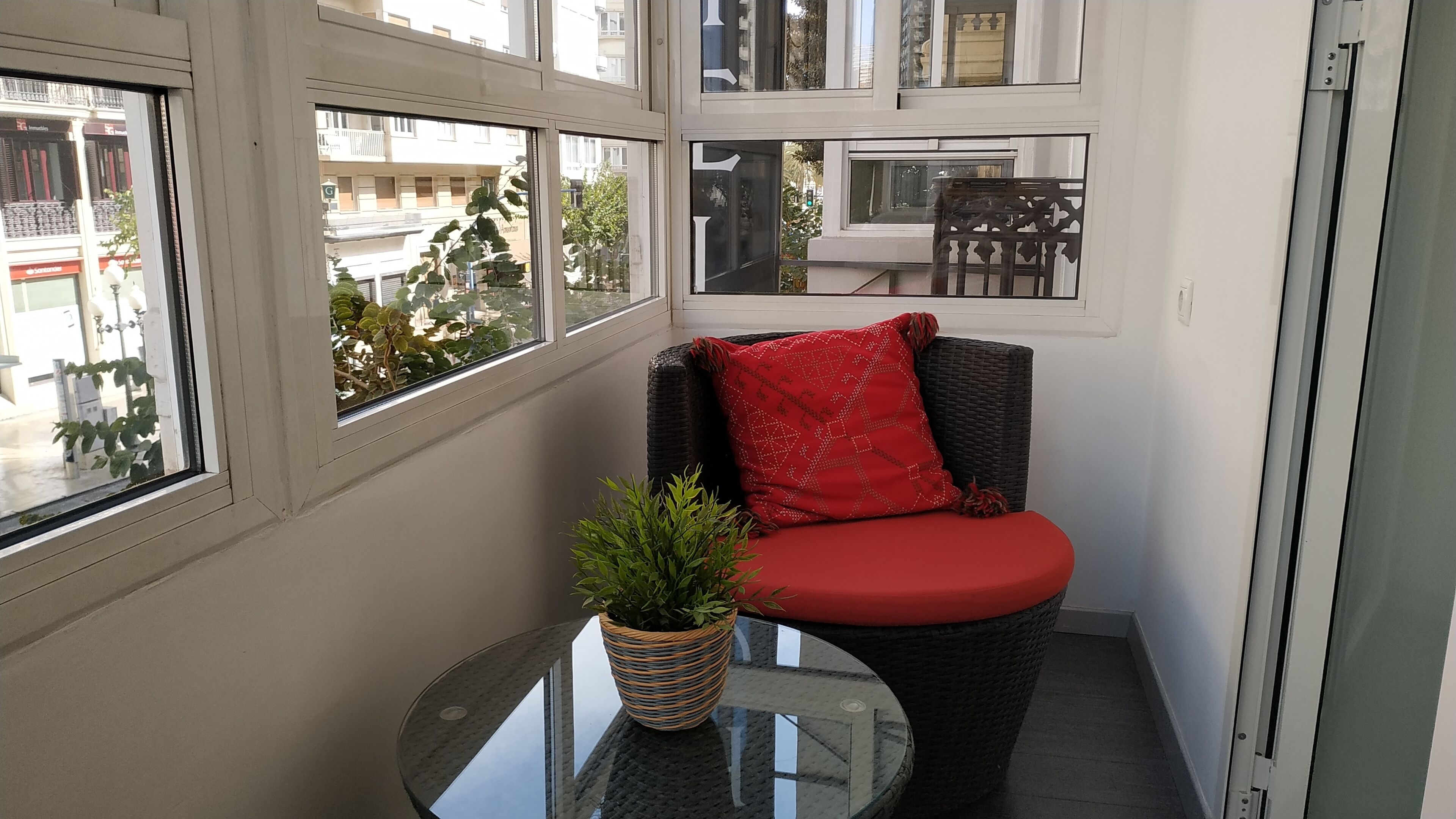 A corner seating area with a wicker chair and a red cushion, a small table with a potted plant, and large windows offering a view of the street.