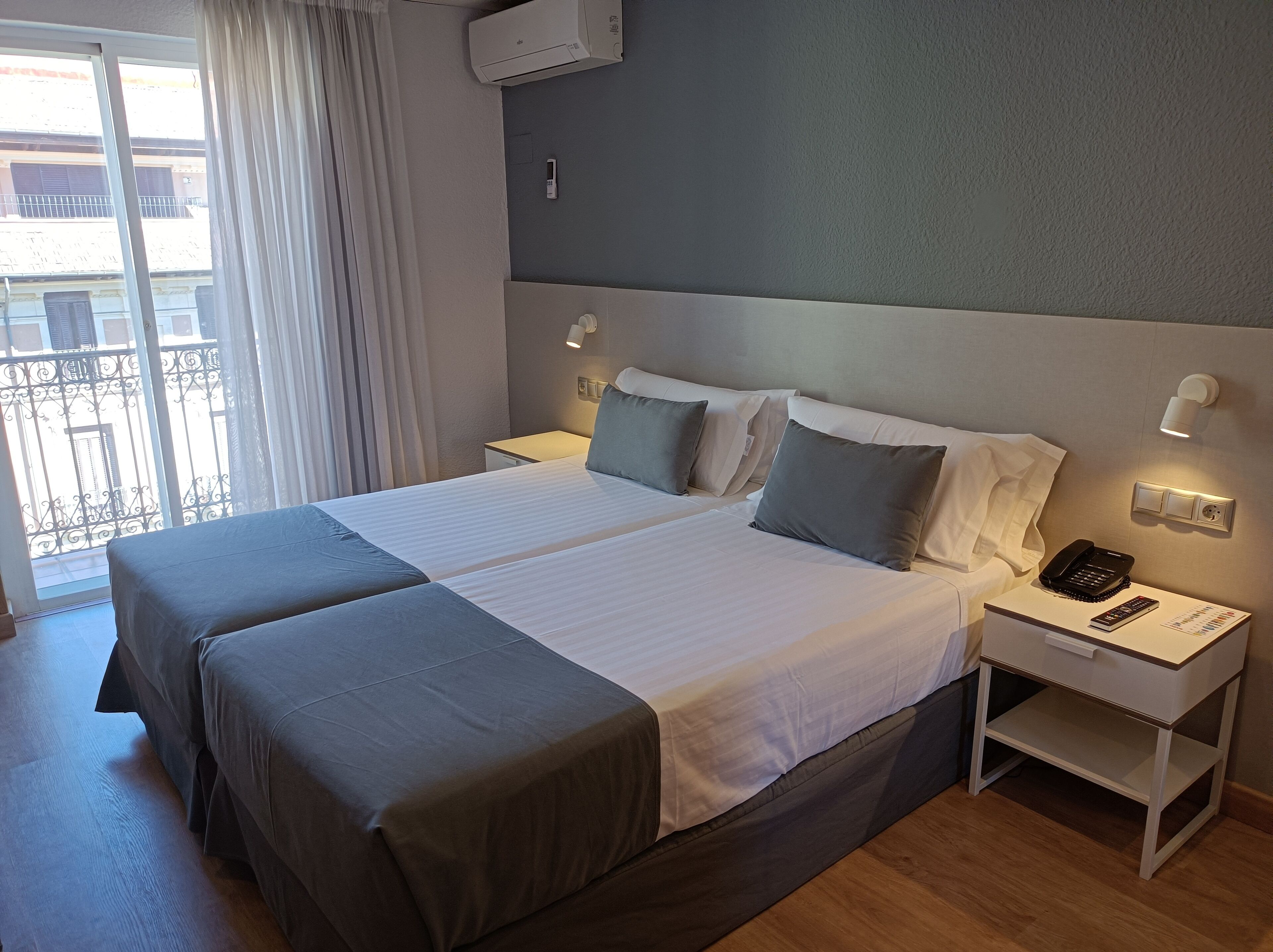 A hotel room with a large bed, two bedside tables, a wall-mounted air conditioner, and a view of the outside balcony.