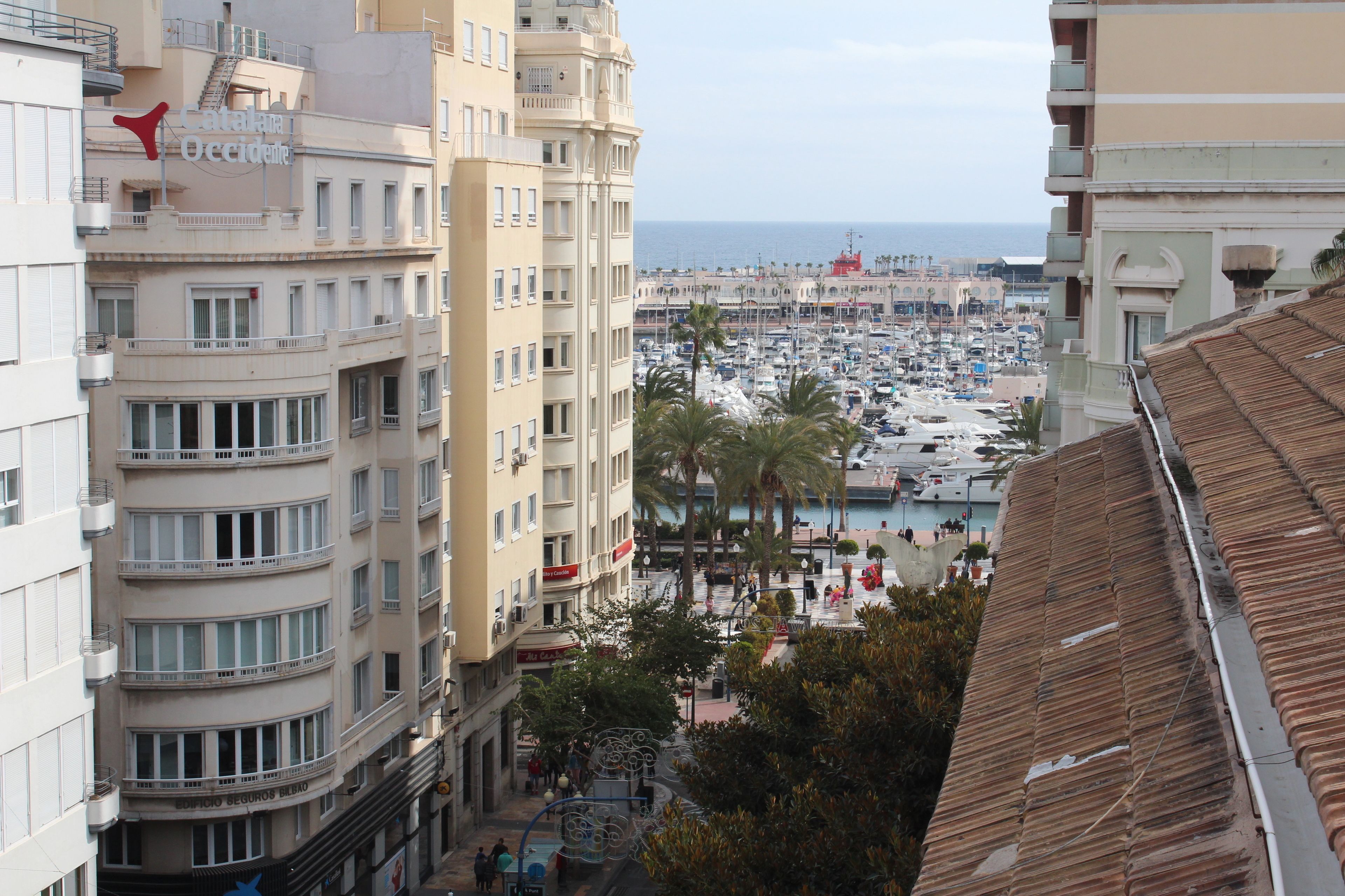 A multi-story hotel with balconies, a marina in the background, and a view of the sea.