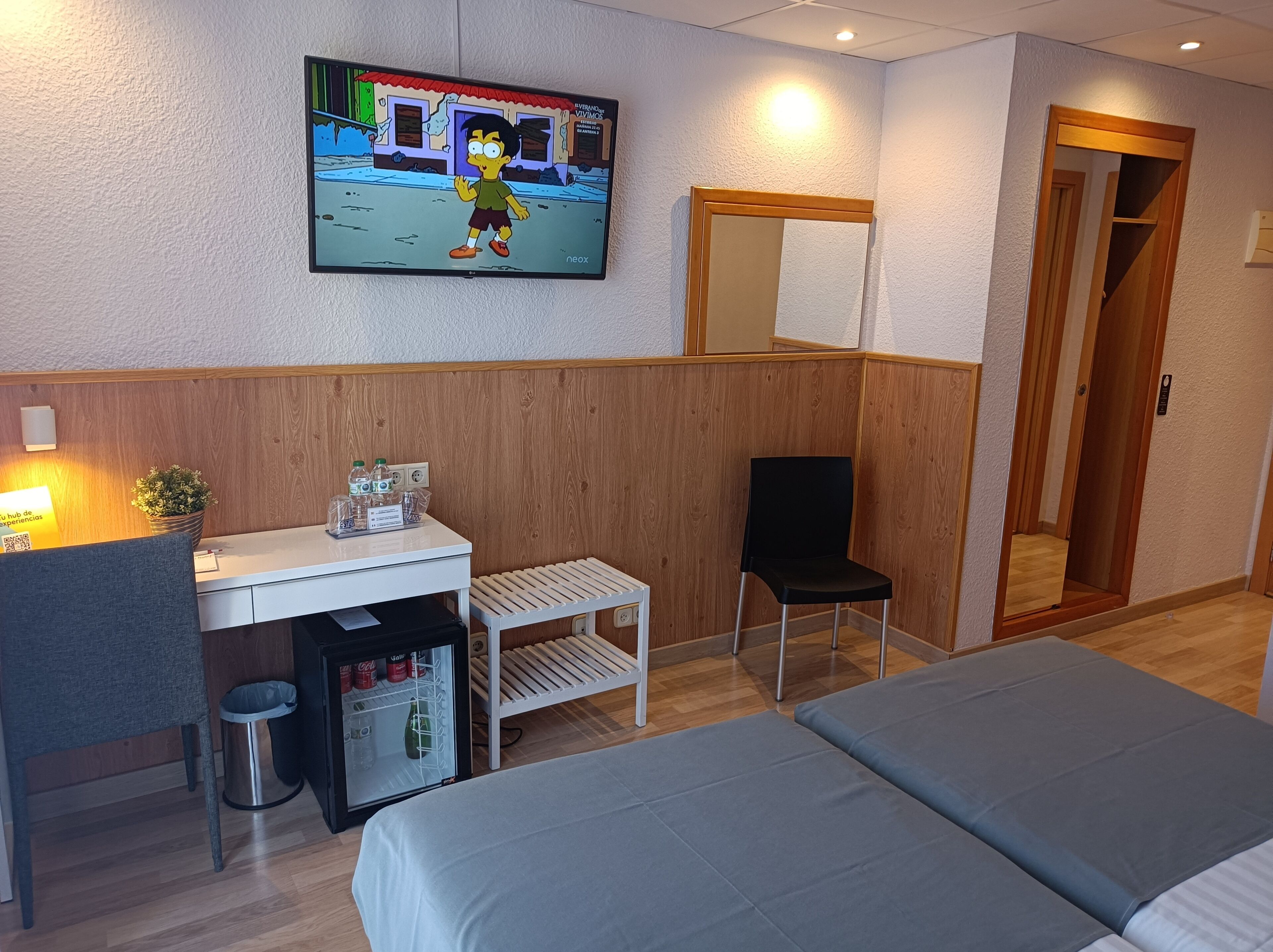 A hotel room with a bed, a desk with a computer, a chair, a small refrigerator, and a TV on the wall.