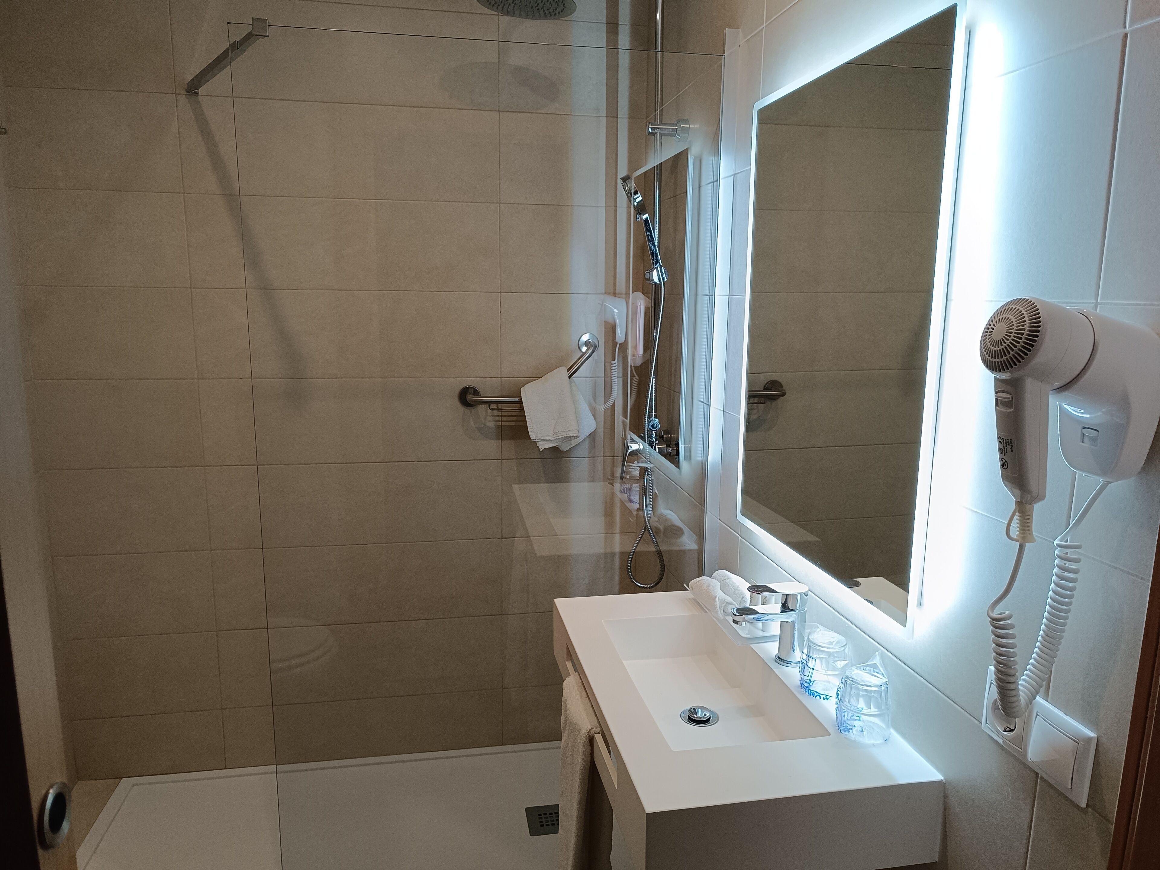 Un bagno moderno con box doccia in vetro, lavandino bianco e specchio sopra, e un asciugacapelli fissato alla parete.