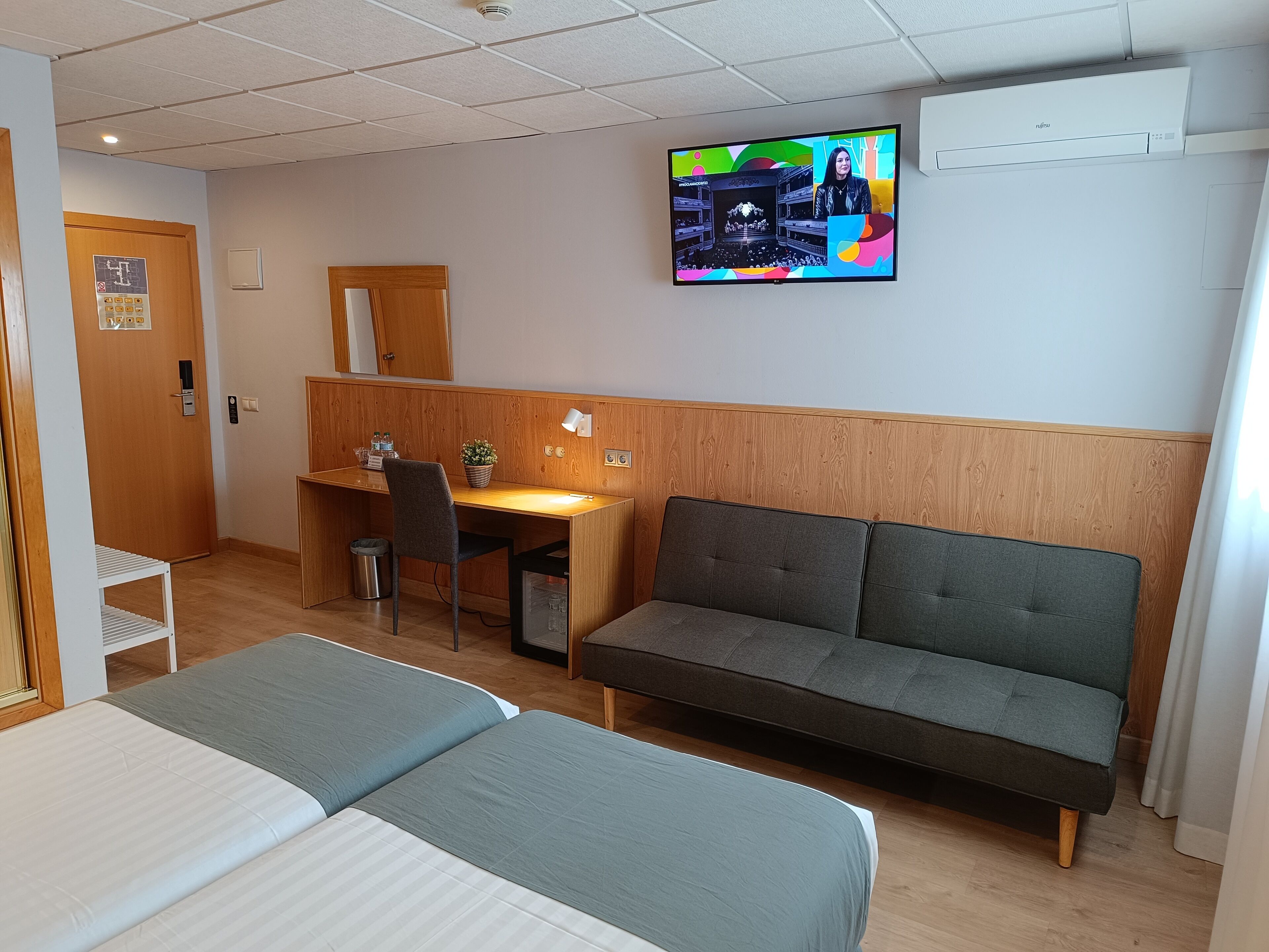 A hotel room with two single beds, a desk with a chair, a sofa, a TV mounted on the wall, and a wooden panel wall.