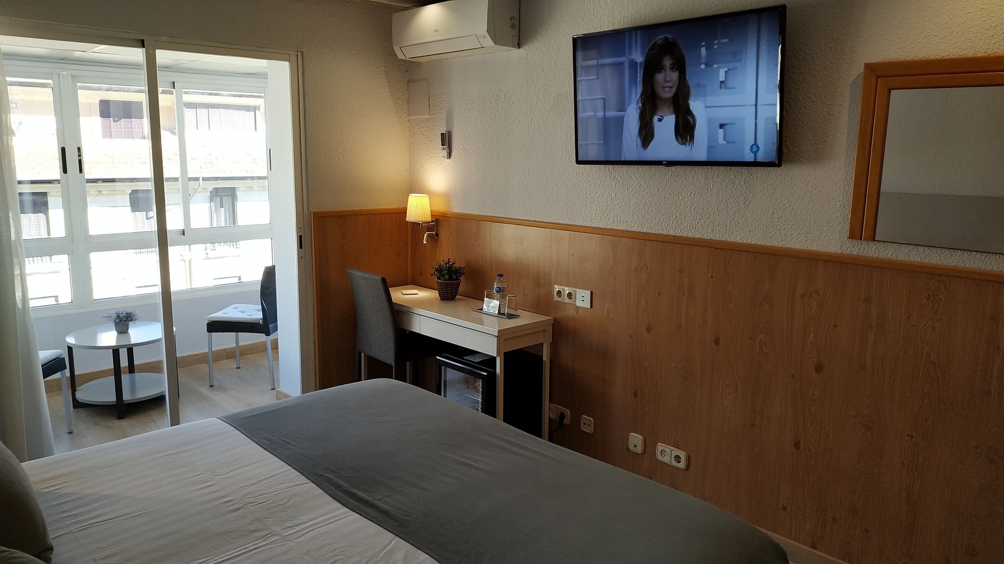 Habitación de hotel con cama, escritorio, silla, lámpara y televisión en la pared.