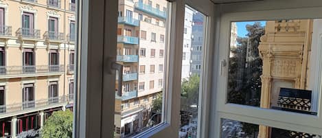 A view from a window overlooking a multi-story building with balconies, a street with cars, and a clear sky.
