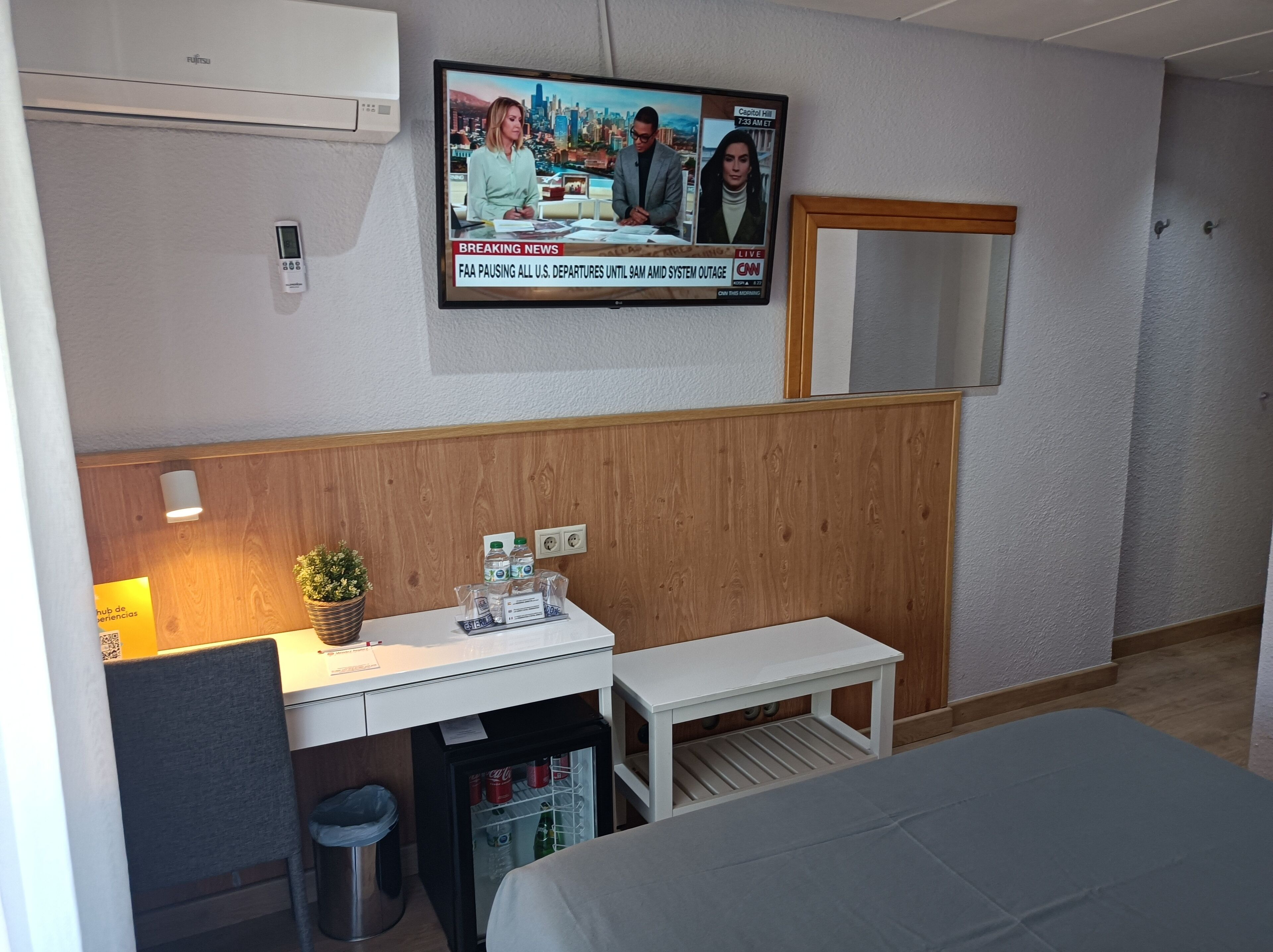 A hotel room with a wooden panel wall, a television mounted above a desk, a small refrigerator, a bed, and a mirror.