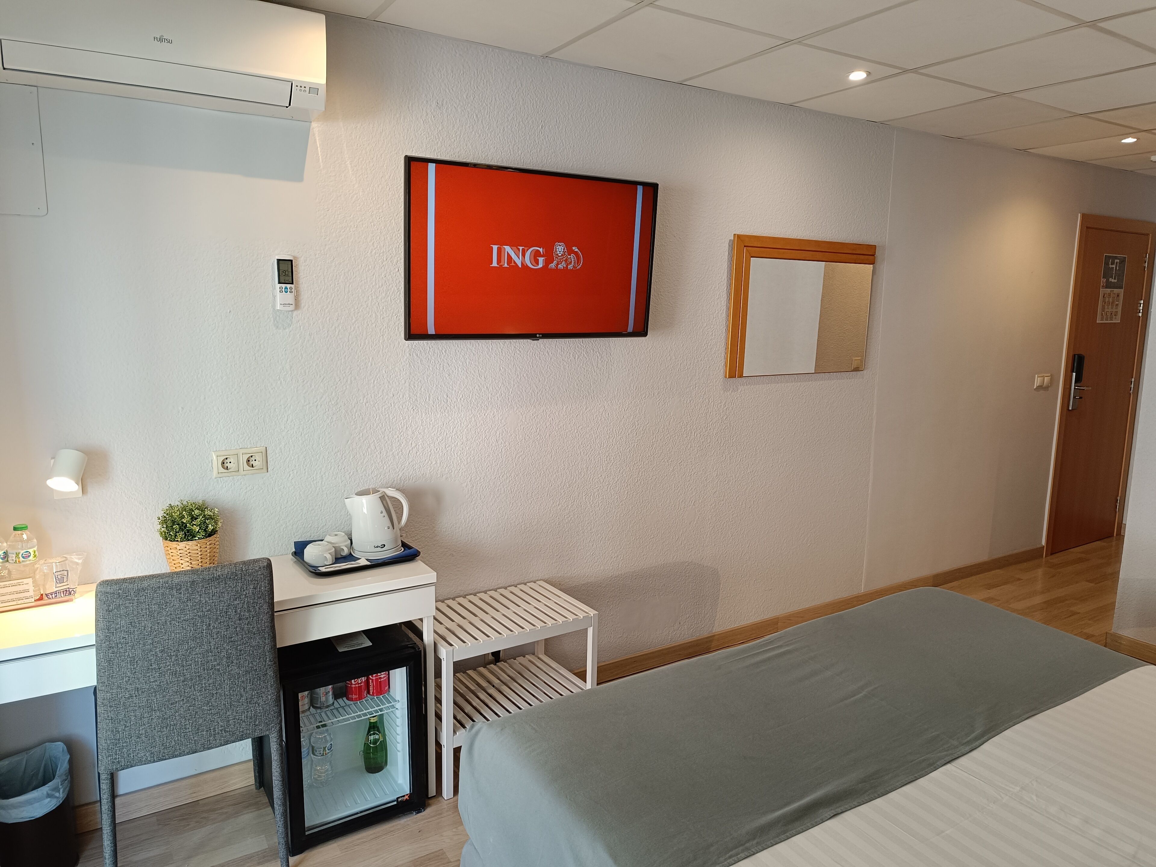 Ein Hotelzimmer mit einem Bett, einem Schreibtisch mit Wasserkocher, einem Minikühlschrank, einem Fernseher und einem Spiegel an der Wand.