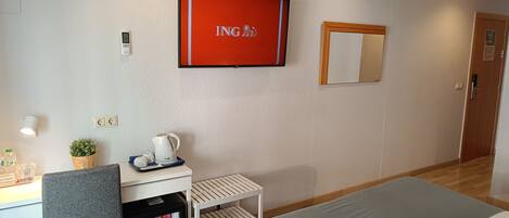 A hotel room with a bed, a desk with a kettle, a mini-fridge, a TV, and a mirror on the wall.