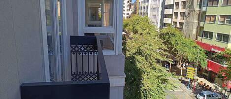 Un edificio de varios pisos con balcones, vista a la calle con coches y peatones, y un restaurante con terraza.