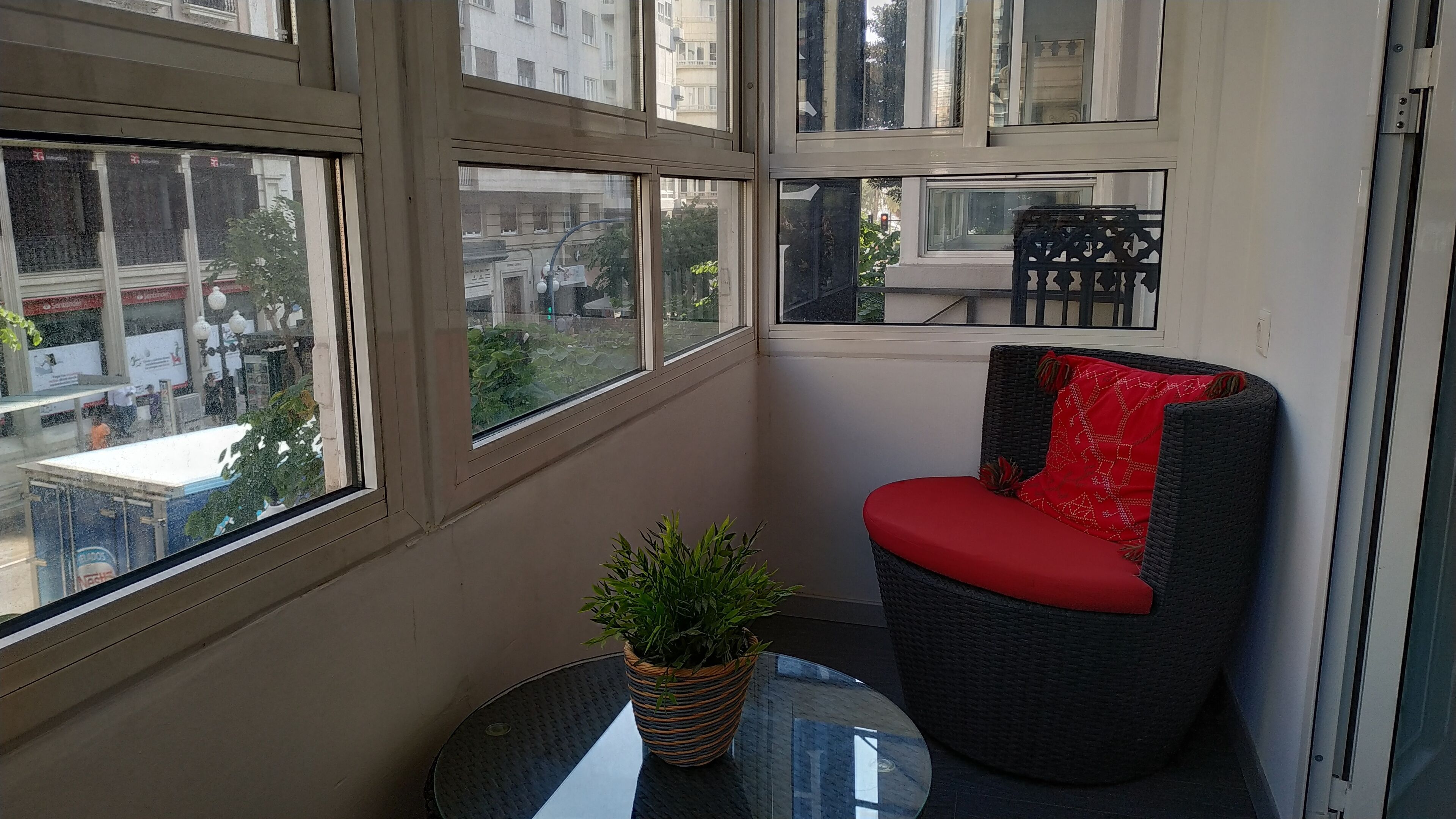 A corner room with a view of the city, a wicker chair with a red cushion, and a potted plant on a small table.