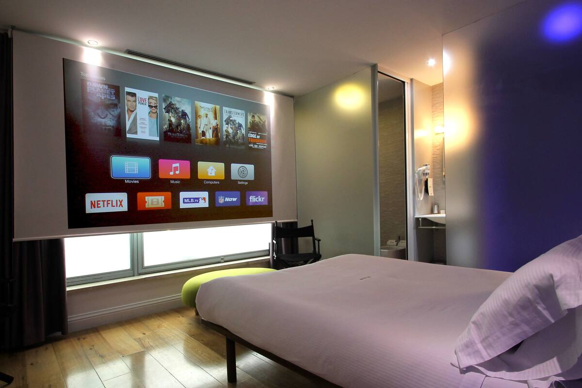 Habitación de hotel con una televisión grande montada en la pared que muestra diferentes servicios de streaming, una cama con almohada, una mesita de noche y una silla.