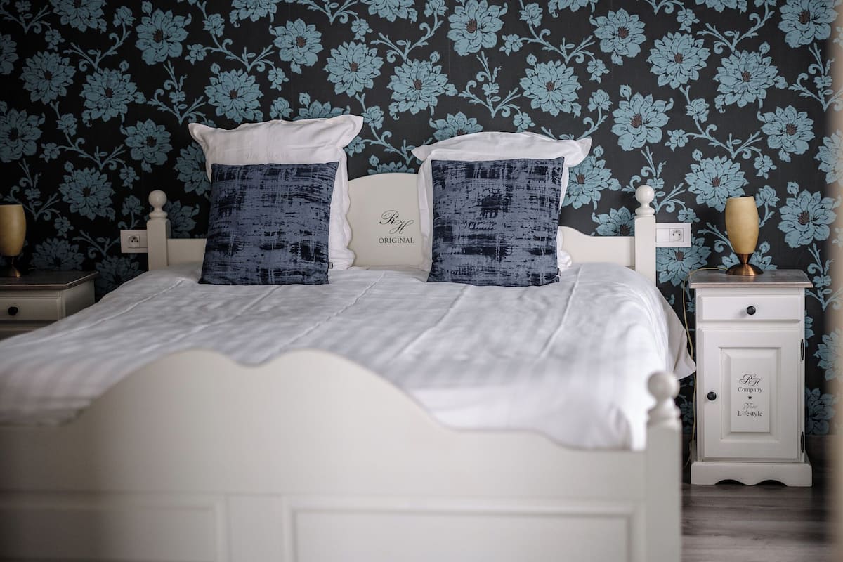 Cama bem arrumada com roupa de cama branca e duas almofadas decorativas, mesa de cabeceira com abajur e parede com papel de parede floral.