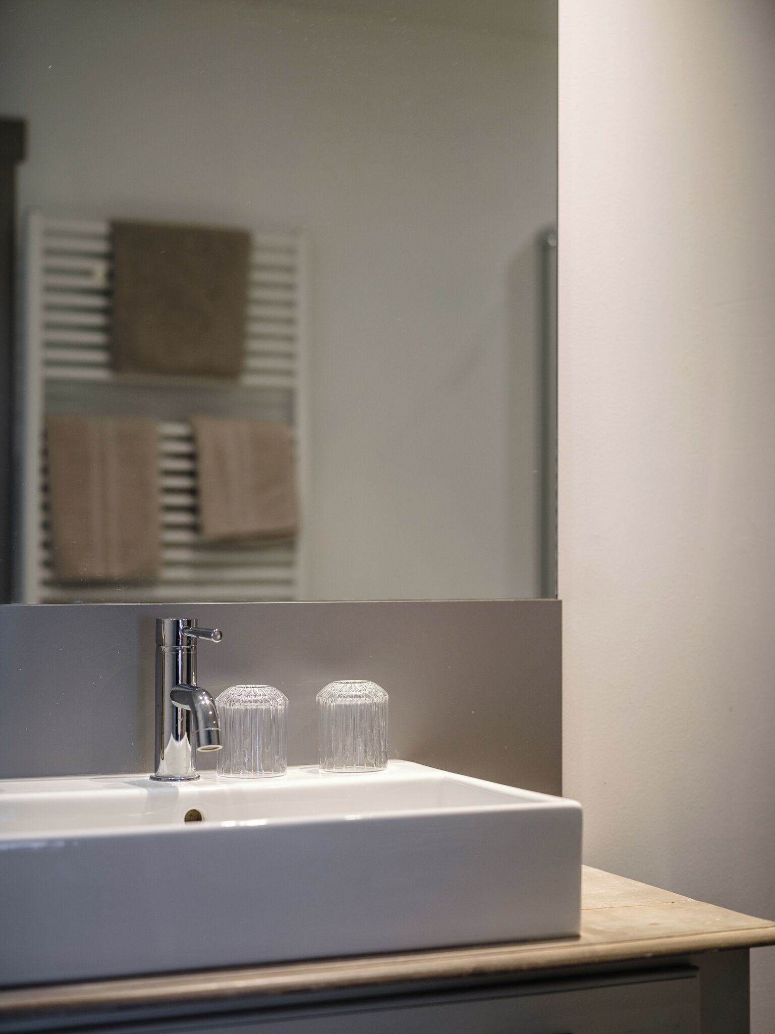 A bathroom with a white sink, two clear glass vases, and a mirror reflecting a towel rack with towels.