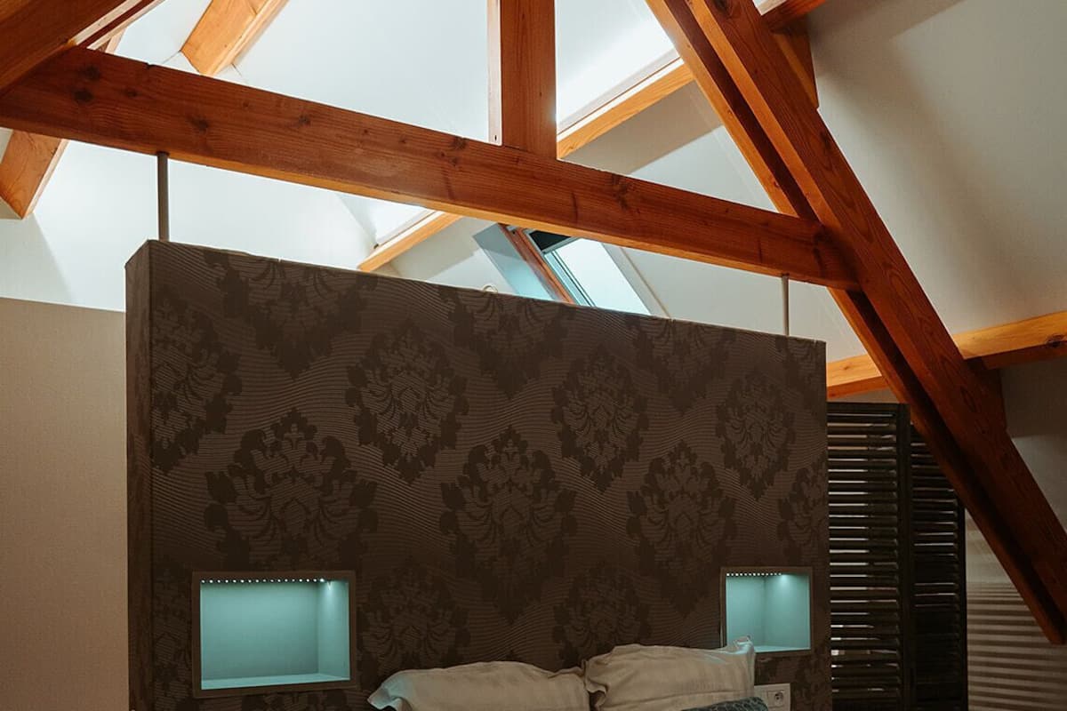 Quarto com teto inclinado, vigas de madeira, cabeceira estampada e duas luminárias de mesa.
