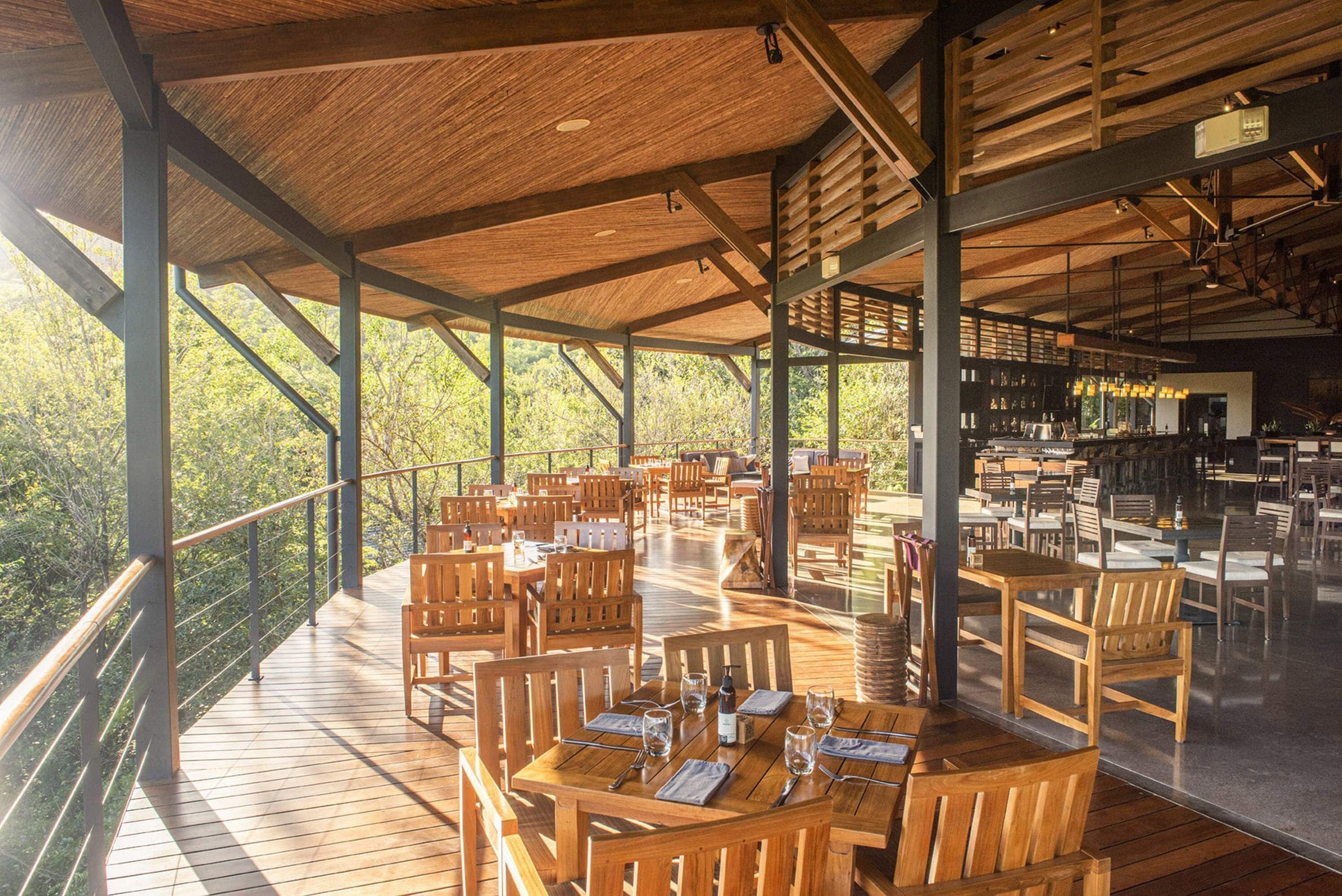 An outdoor dining area with wooden tables and chairs, surrounded by glass railings, and overlooking a forested area.