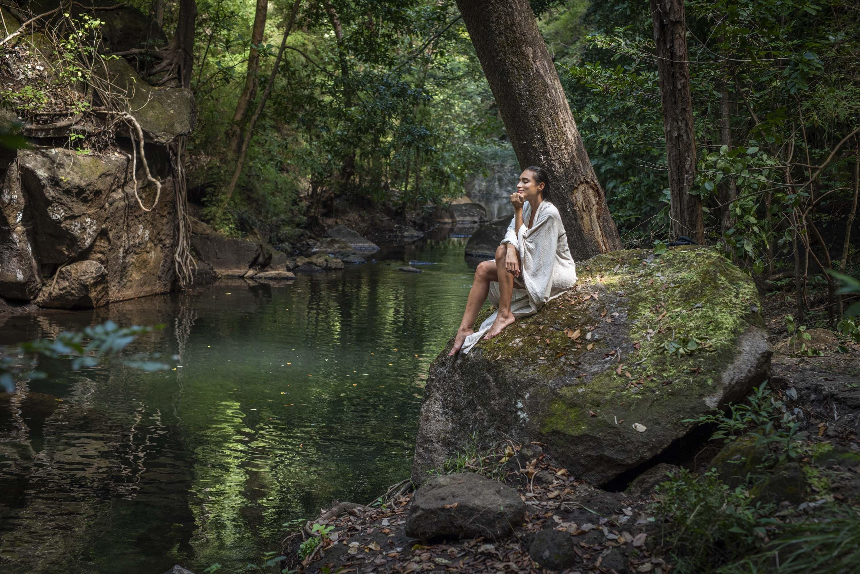 A person sitting on a moss-covered rock by a calm river surrounded by dense forest.