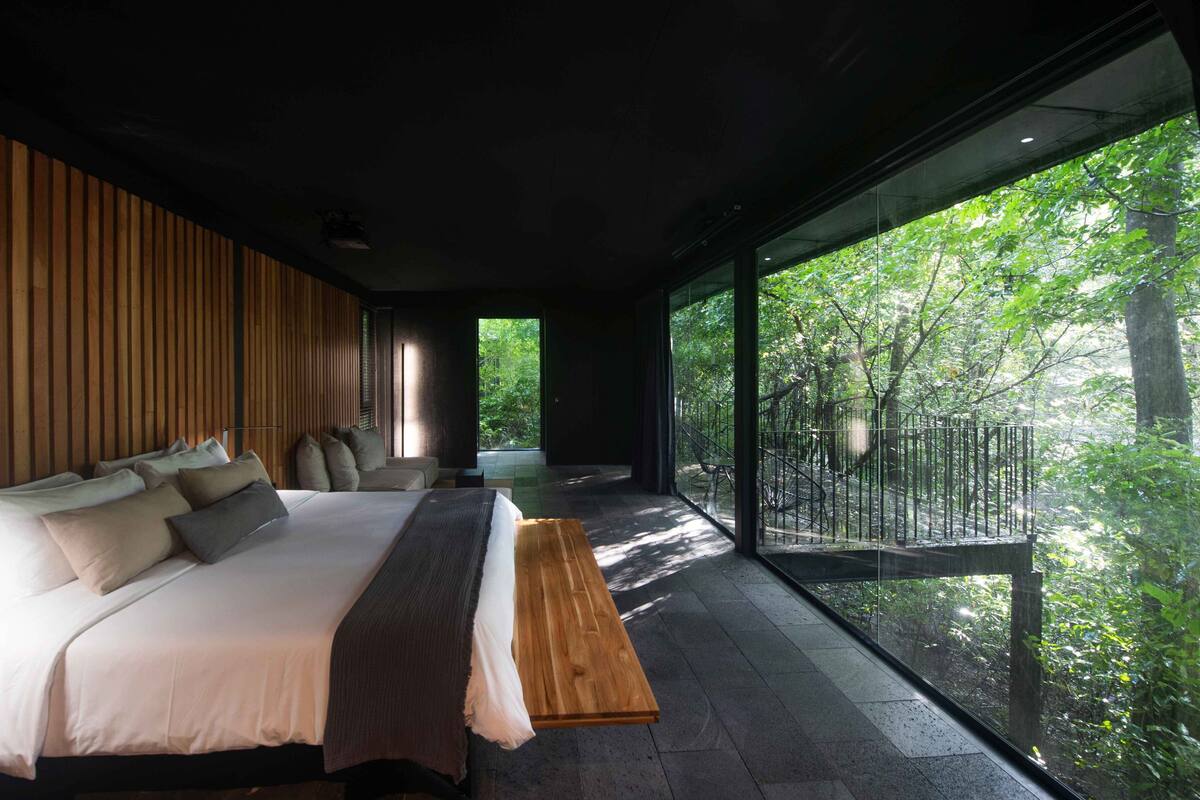 A modern bedroom with a large bed, a wooden bench, and a view of a forest through large windows.