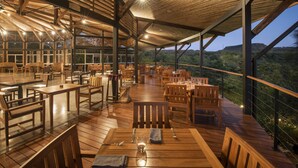 An outdoor dining area with wooden tables and chairs, large windows, and a view of the surrounding landscape. - Rio Perdido Hotel & Thermal River (Fortuna)