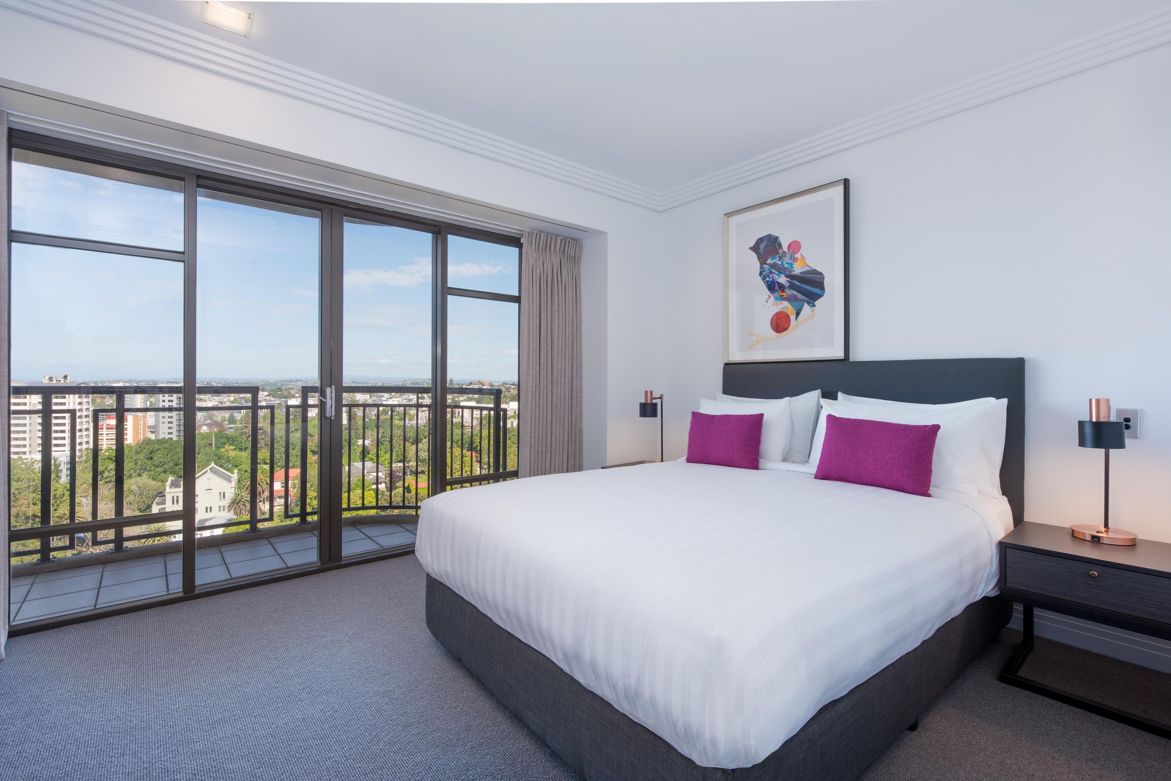 A hotel room with a large bed, a nightstand, a framed picture, and a view through floor-to-ceiling windows.