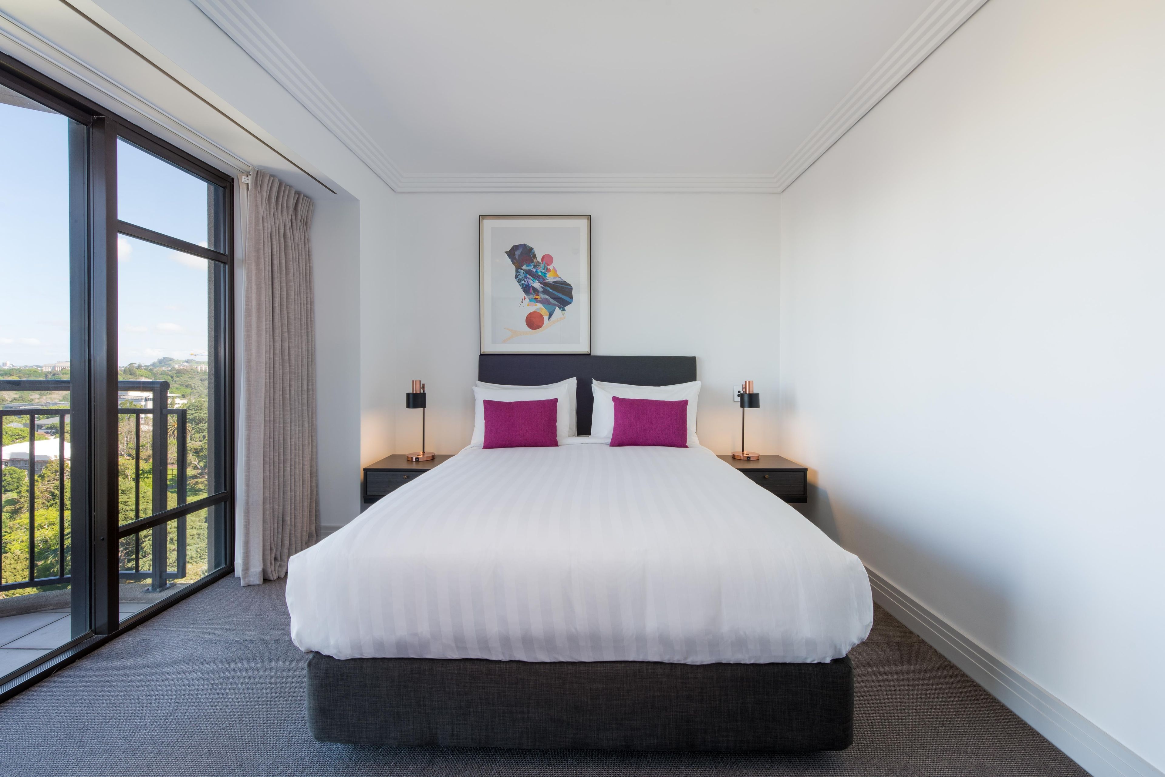A hotel room with a large bed, two nightstands, a framed picture on the wall, and a view through a large window.