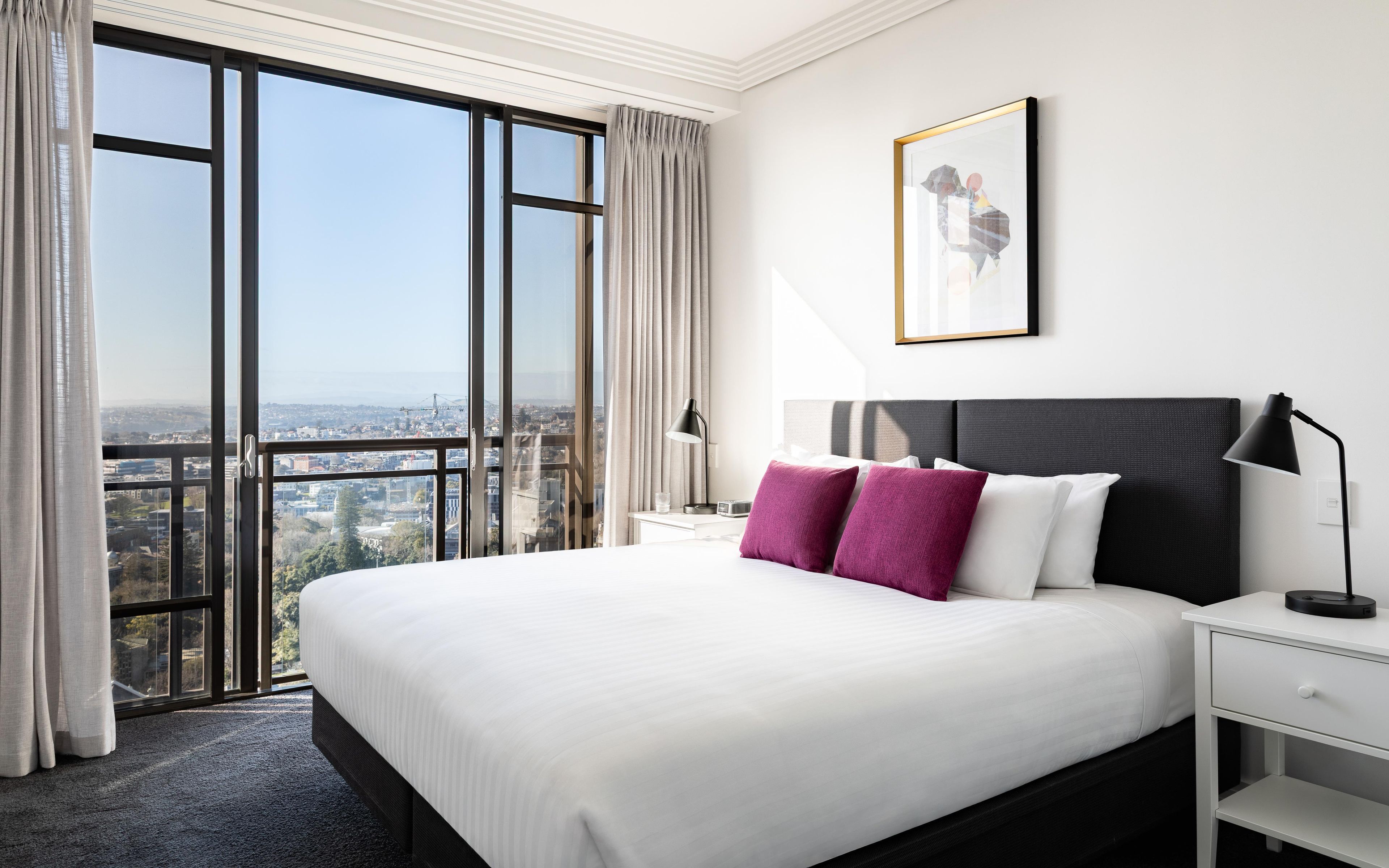 A hotel room with a large bed, a nightstand, a desk, and a view of the city through floor-to-ceiling windows.