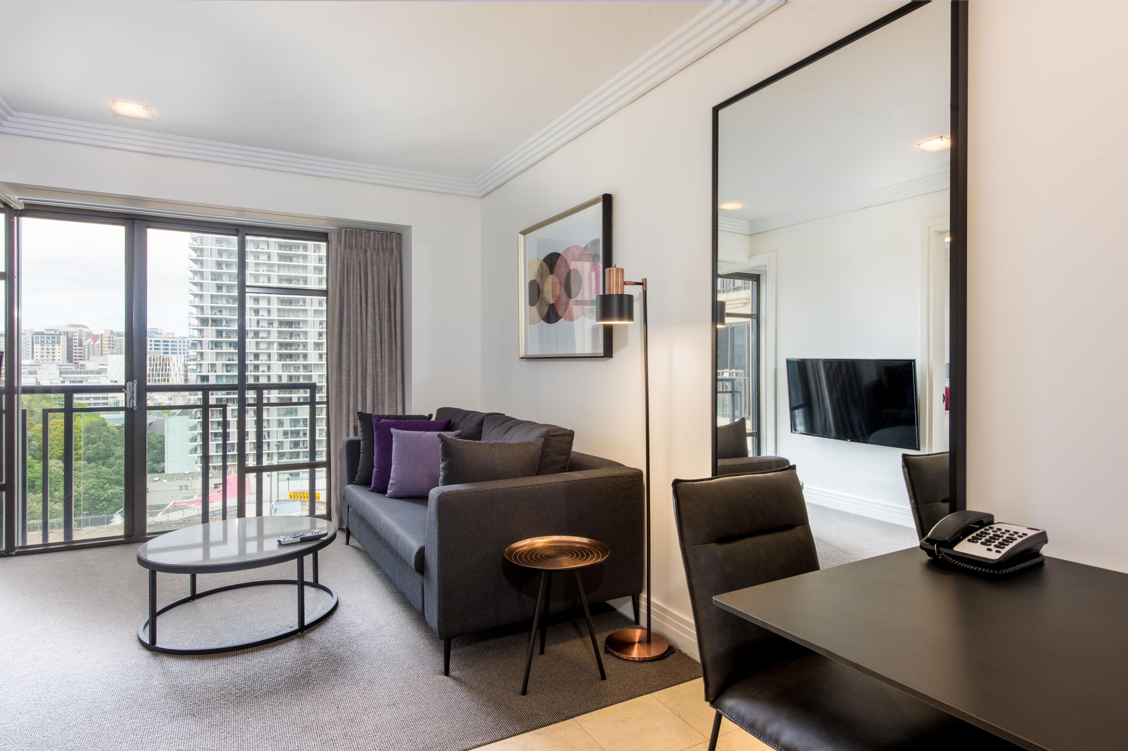 A modern hotel room with a large window, a sofa, a round coffee table, a desk with a telephone, and a floor lamp.