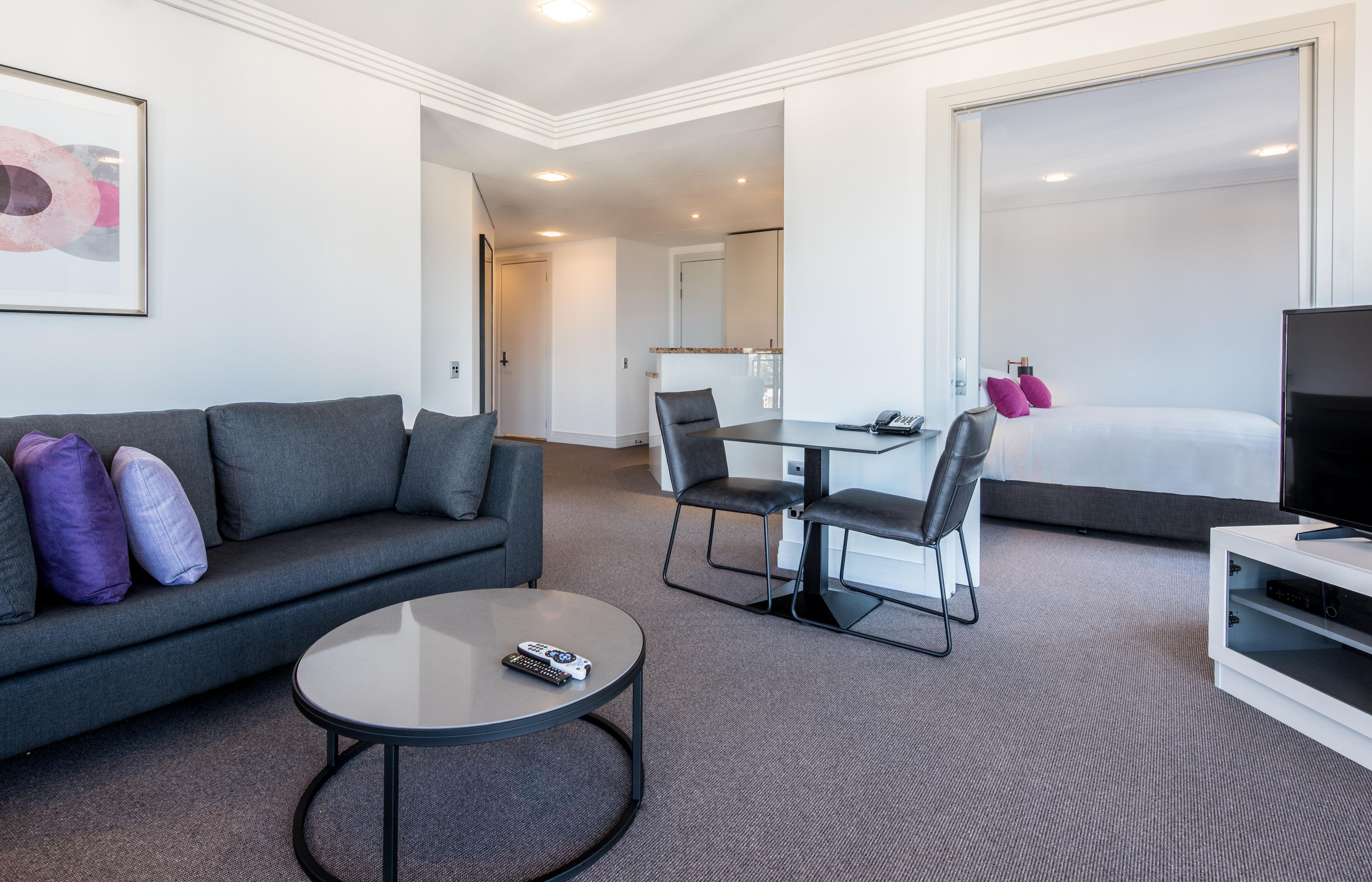 A modern hotel room with a gray sofa, a round coffee table, a dining table with chairs, a TV stand, and a large bed.