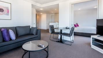 A modern hotel room with a gray sofa, a round coffee table, a dining table with chairs, a TV stand, and a large bed.