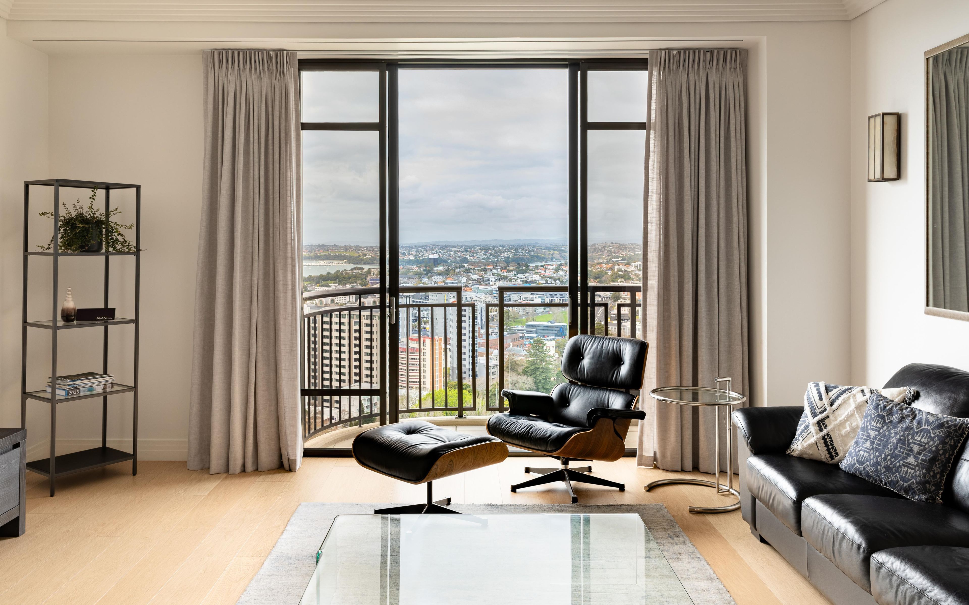 A modern living room with a black leather sofa, a glass coffee table, and a view of a cityscape through large windows.