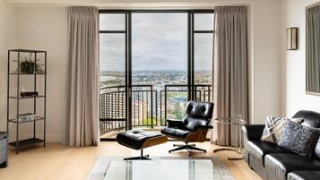 A modern living room with a black leather sofa, a glass coffee table, and a view of a cityscape through large windows.