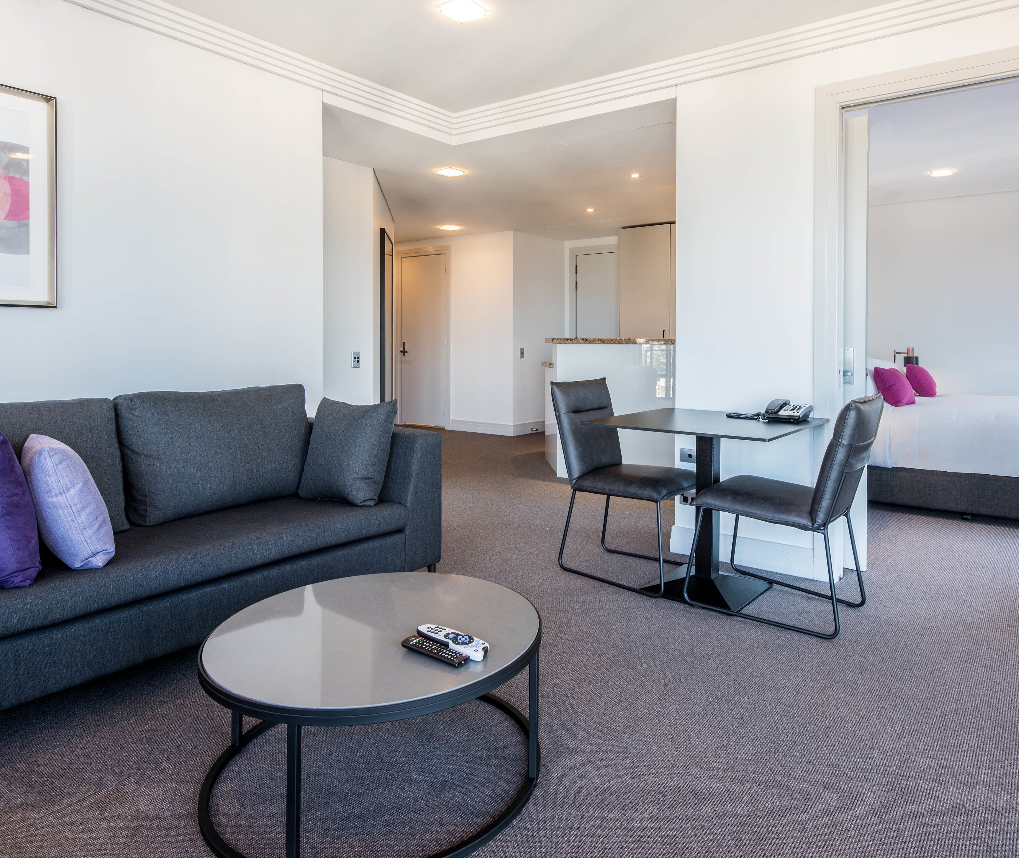 Una habitación de hotel moderna con un sofá gris, una mesa redonda y un comedor con una mesa y sillas.