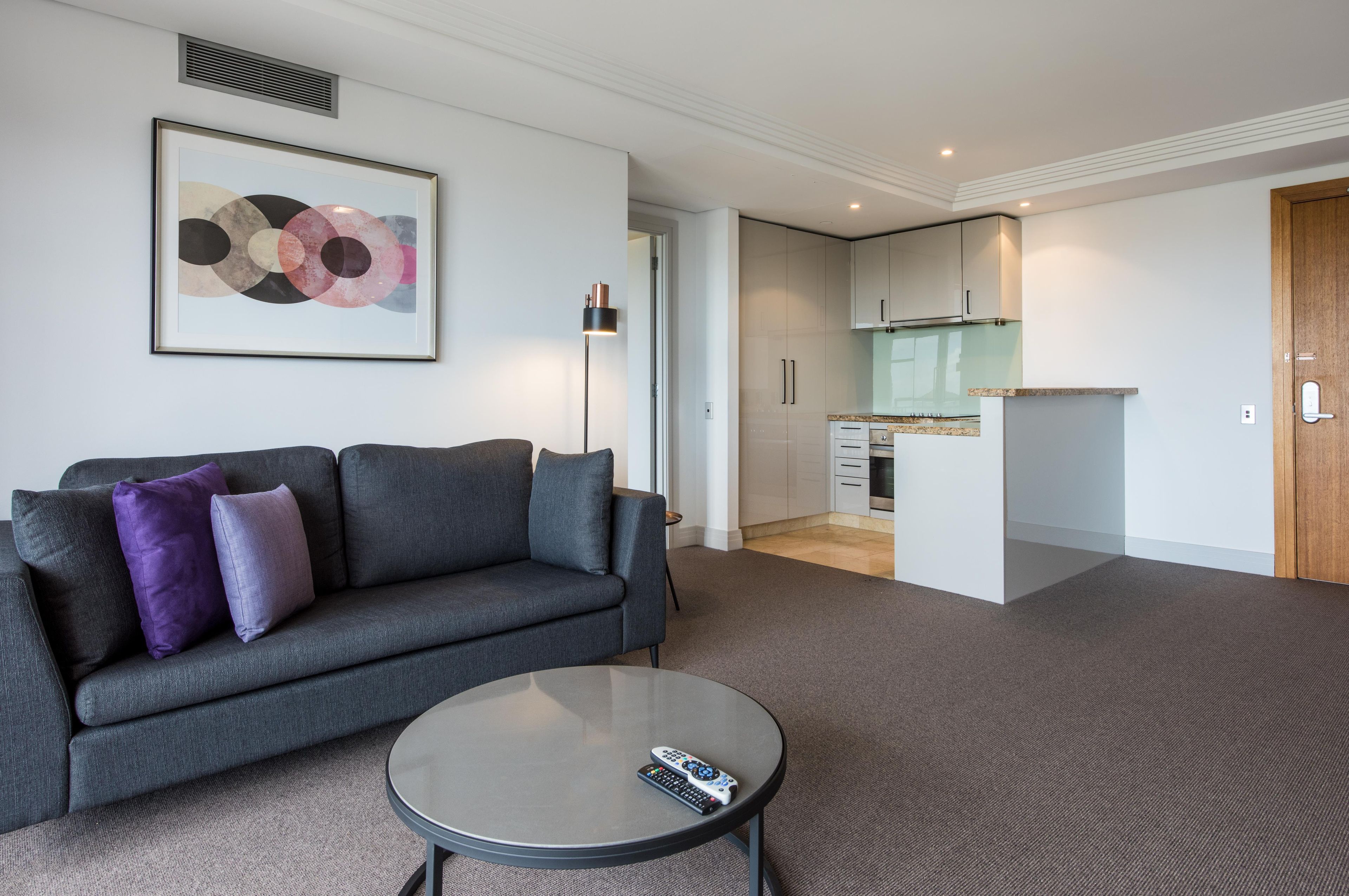 A modern living room with a grey sofa, a round coffee table, and a remote control. There is a kitchen in the background with white cabinets and a glass backsplash.