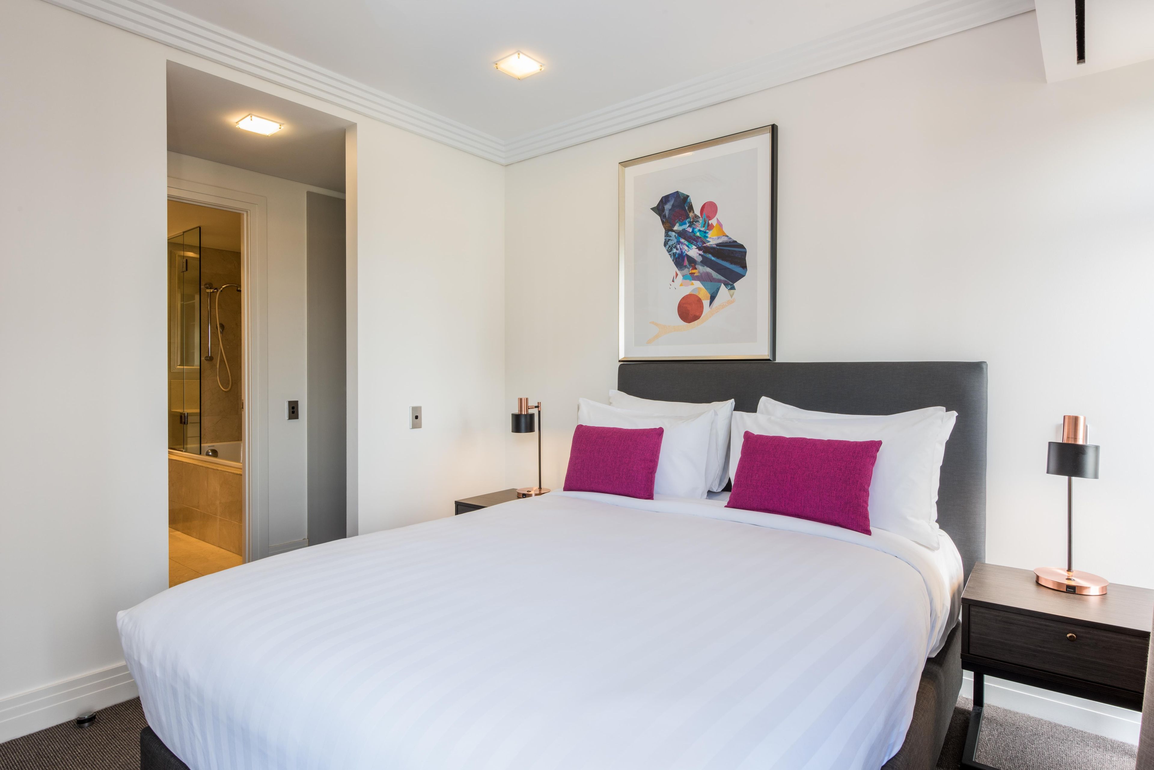 A hotel room with a large bed, a nightstand, a wall-mounted artwork, and a door leading to a bathroom.