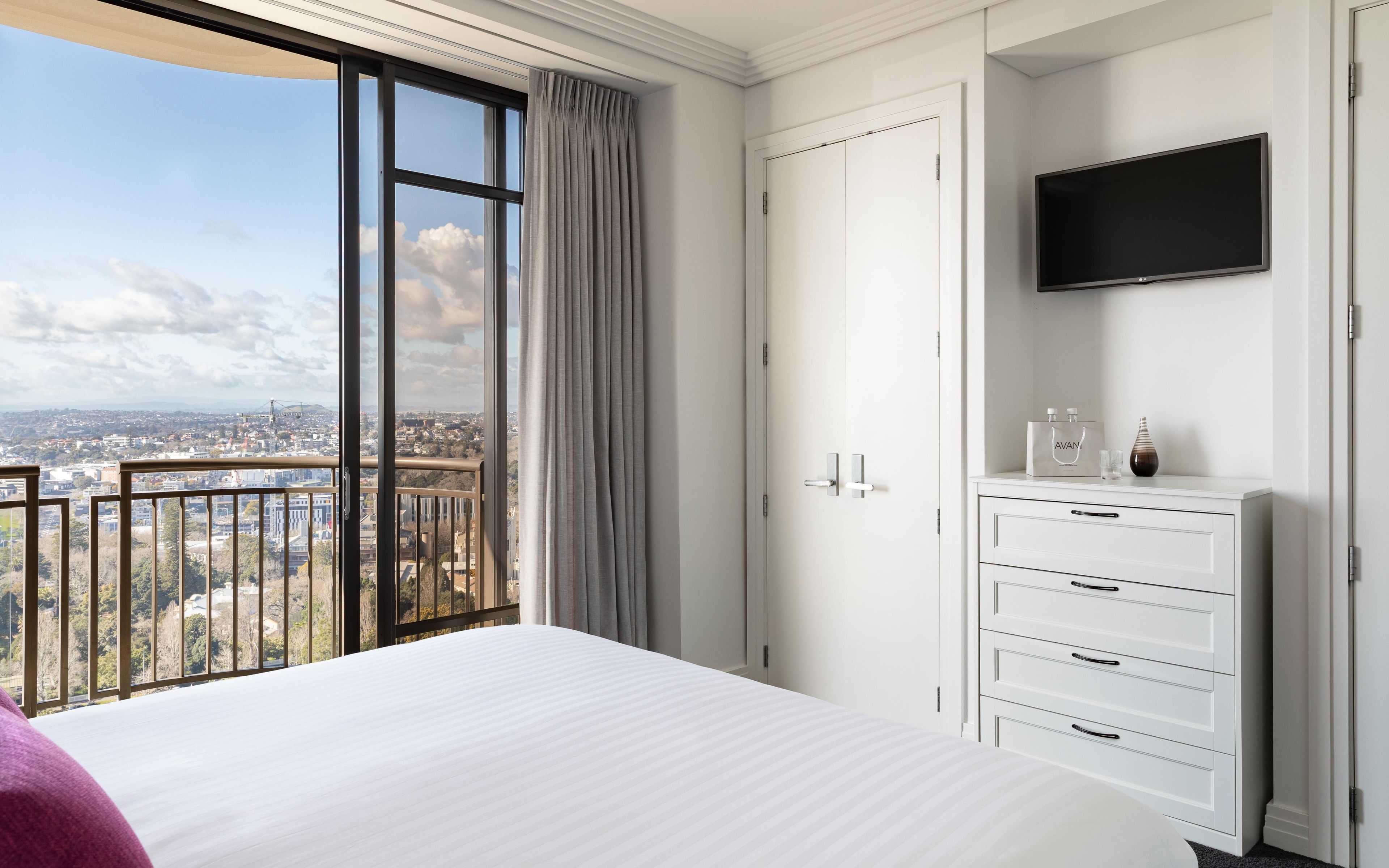 A hotel room with a large window, a bed, a dresser, and a flat-screen TV.