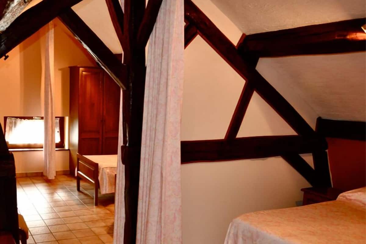 A bedroom with a sloped ceiling, wooden beams, a bed, a chair, and a window with curtains.