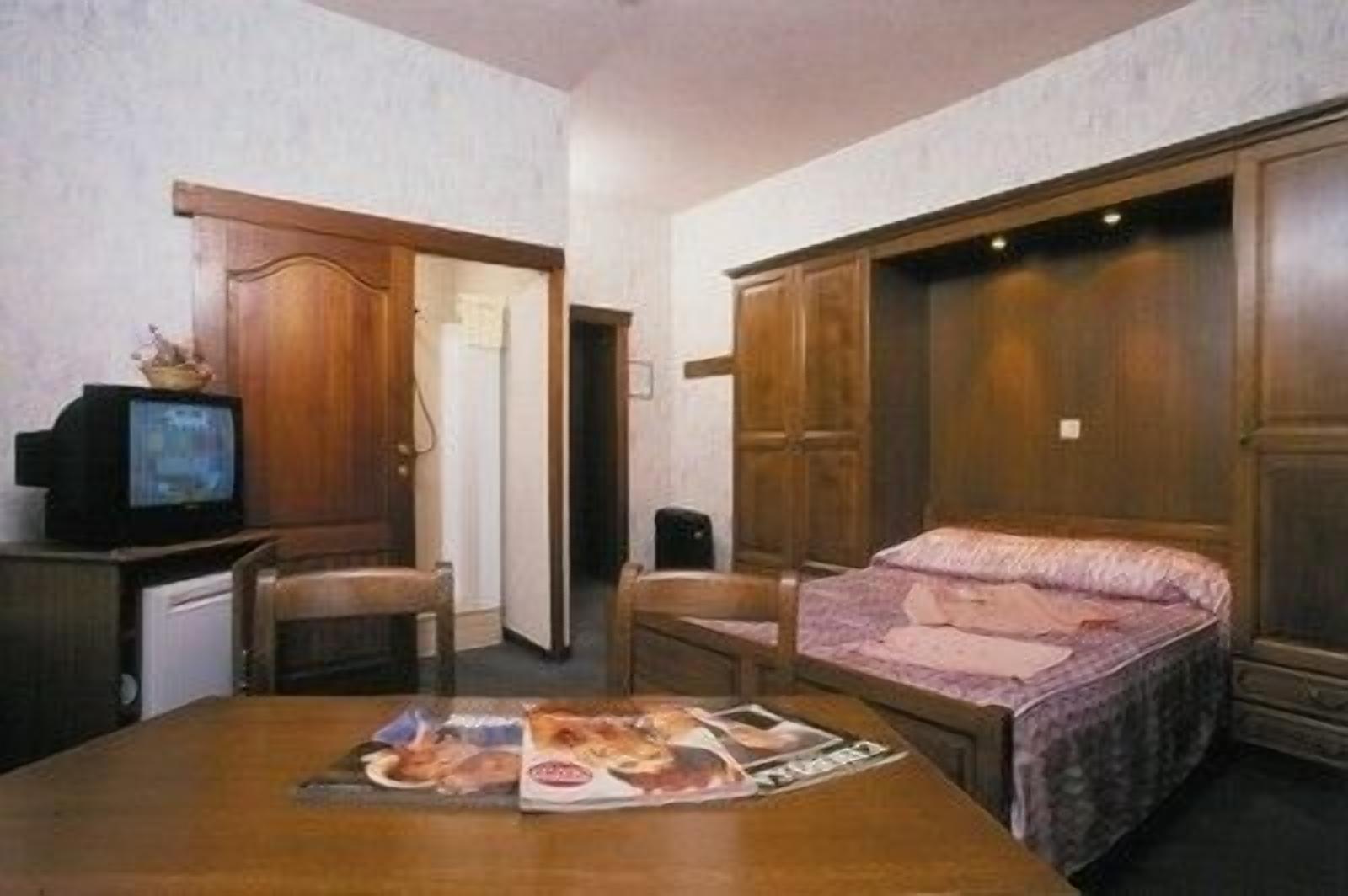 A room with a bed, a desk with a computer, and a television. There are wooden doors and a wardrobe.