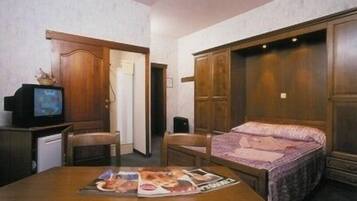A room with a bed, a desk with a computer, and a television. There are wooden doors and a wardrobe.
