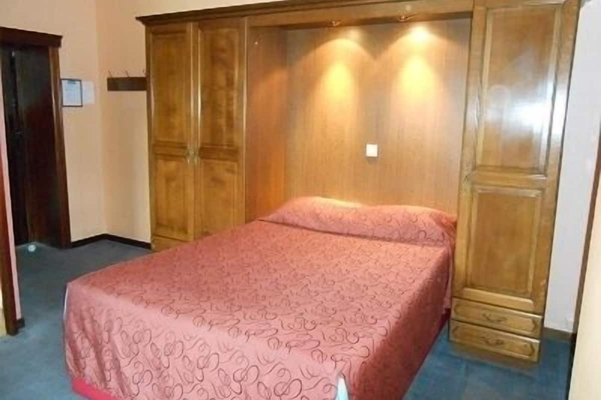 A bedroom with a large wooden wardrobe, a bed with a patterned bedspread, and a blue carpet.