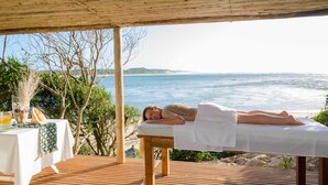 A wooden deck with a massage table, a table with a glass of drink and a decorative item, and a view of the sea.