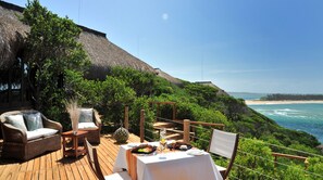 A wooden deck with wicker furniture and a table set for dining, overlooking a beach and ocean.