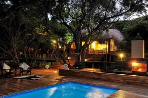 A wooden deck with a pool, outdoor seating, and a thatched-roof structure.