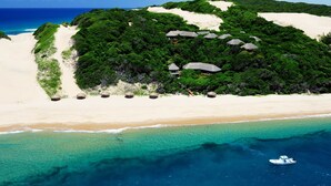 An aerial view of a beach resort with thatched-roof huts, a sandy beach, and clear turquoise waters.