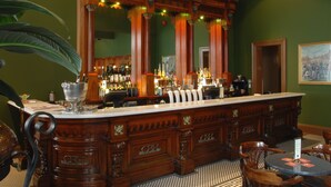 A bar area with a wooden bar counter, bar stools, and a large mirror. There are bottles and glasses on the counter, and a potted plant is visible.