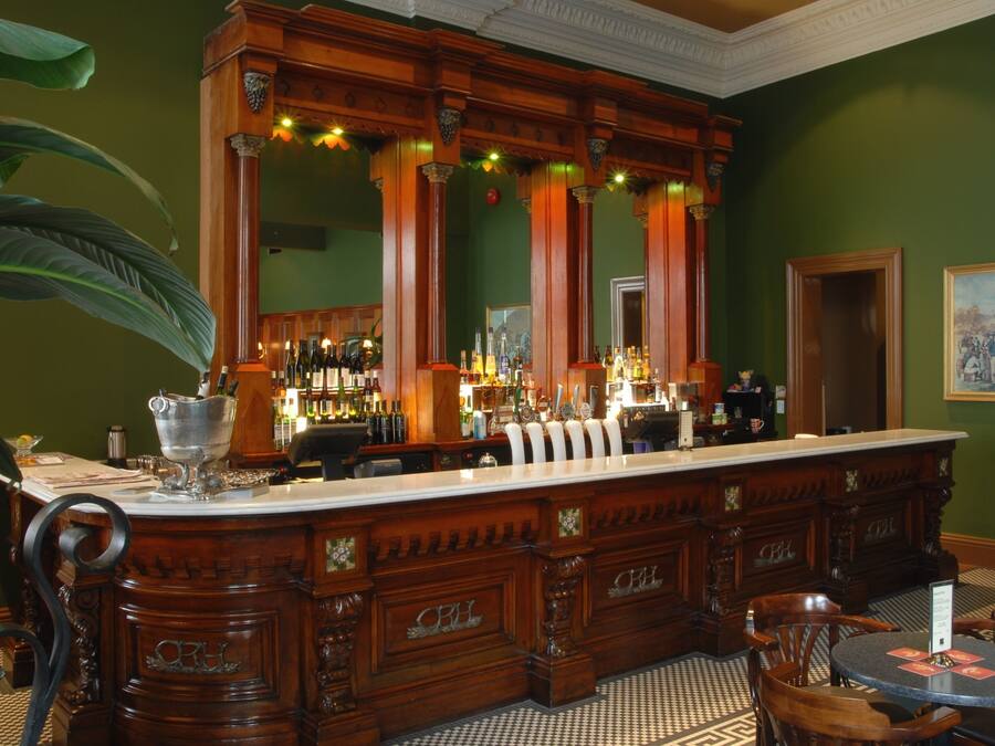 A bar area with a wooden bar counter, bar stools, and a large mirror. There are bottles and glasses on the counter, and a potted plant is visible.