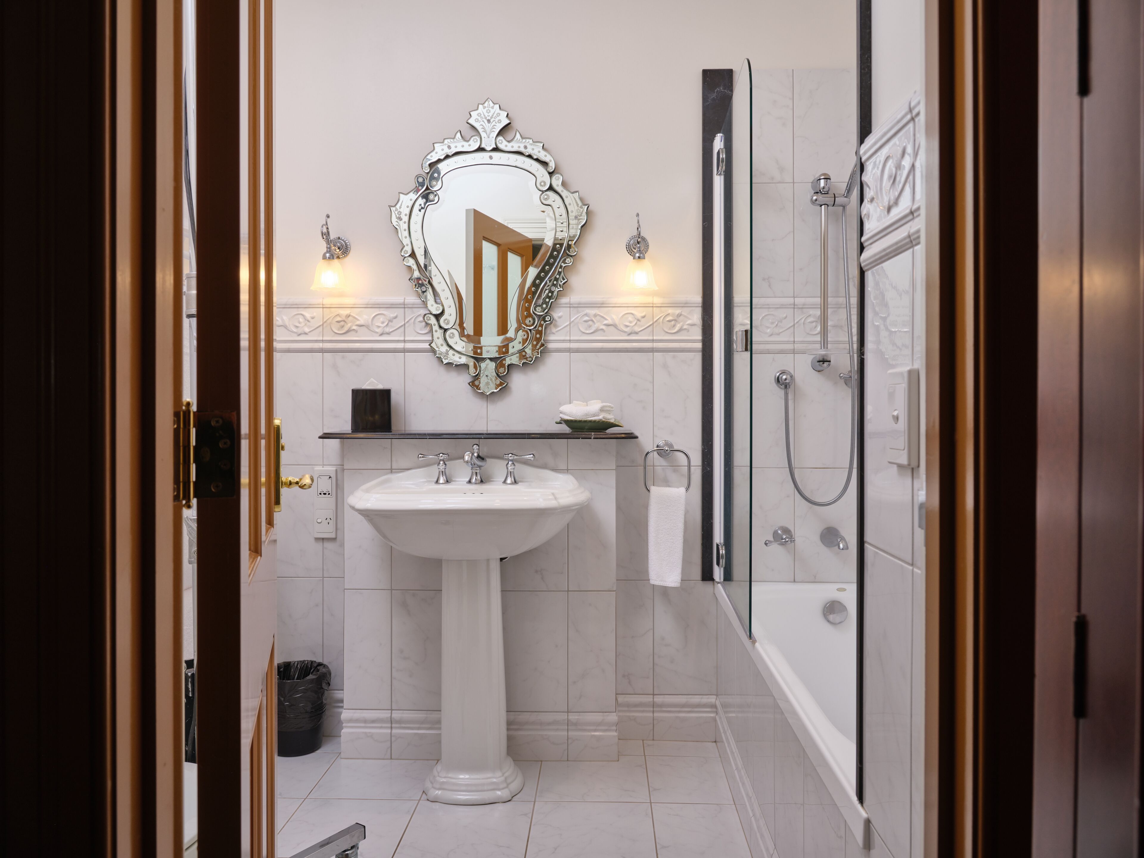 Un baño con lavamanos pedestal, un espejo grande y ornamentado, ducha con cerramiento de vidrio y suelo de baldosas blancas.