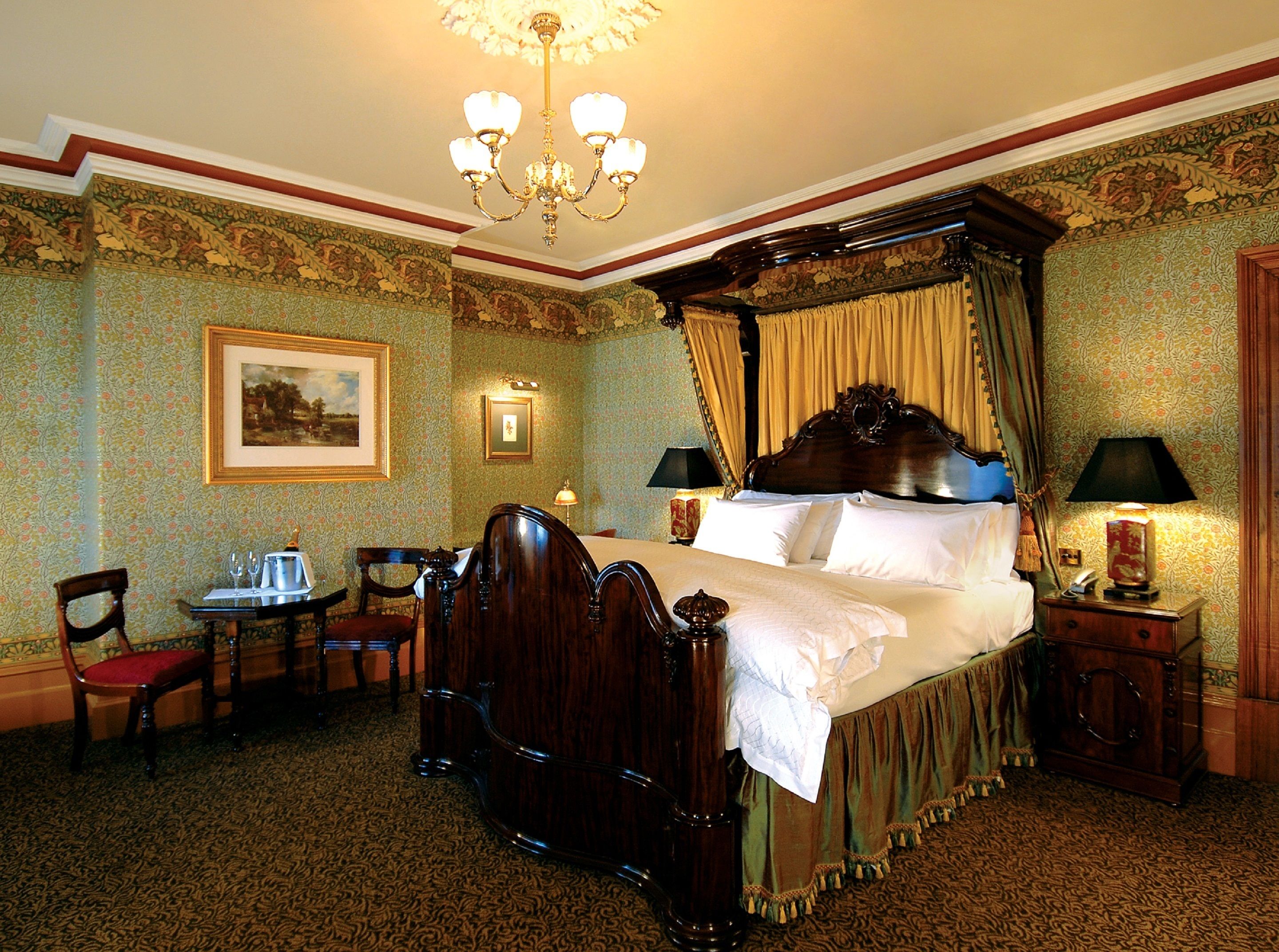 A traditional four-poster bed with a canopy, a chandelier, a desk with a chair, and a telephone on a bedside table.