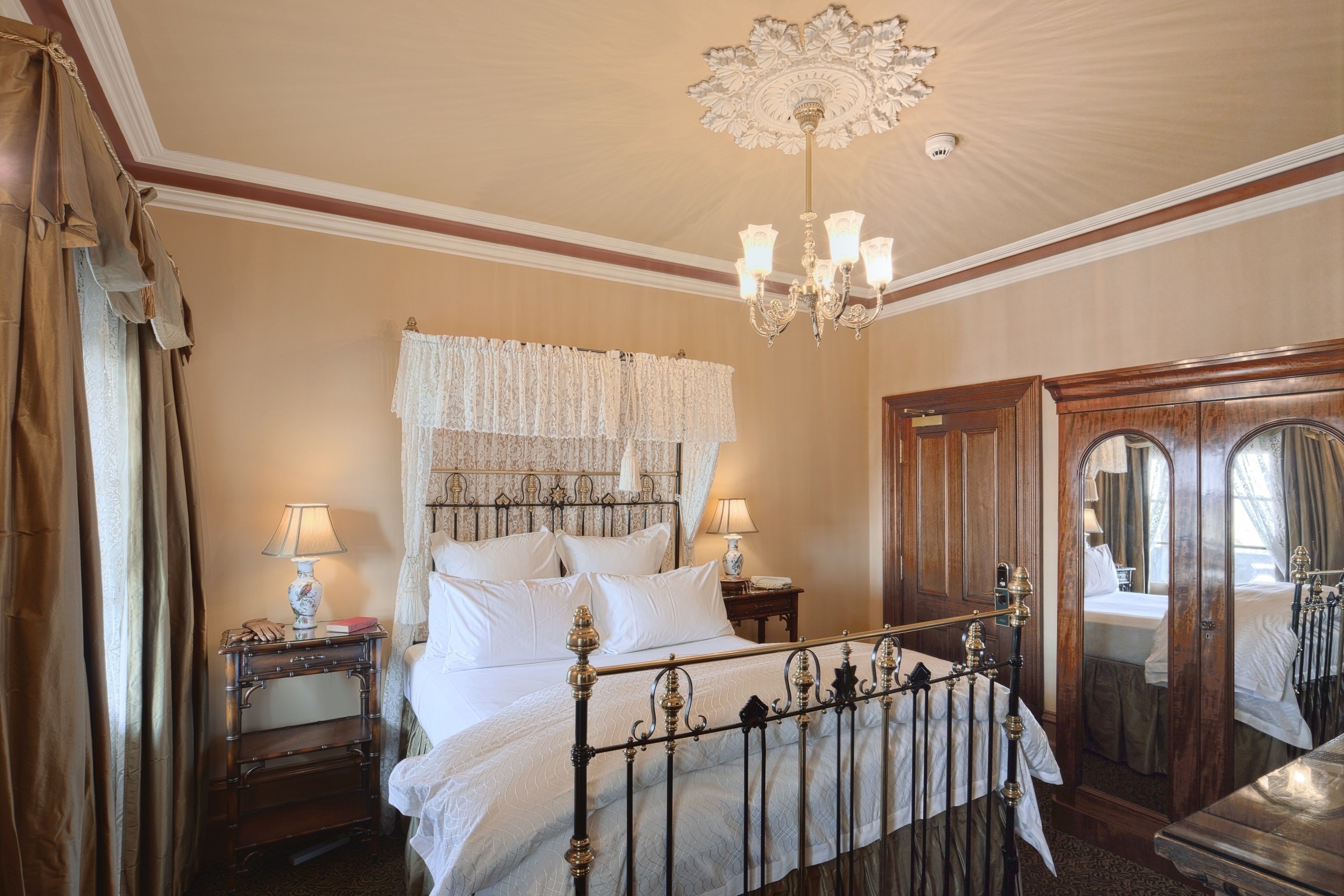 A classic hotel room with a large bed, ornate ceiling, and a chandelier.