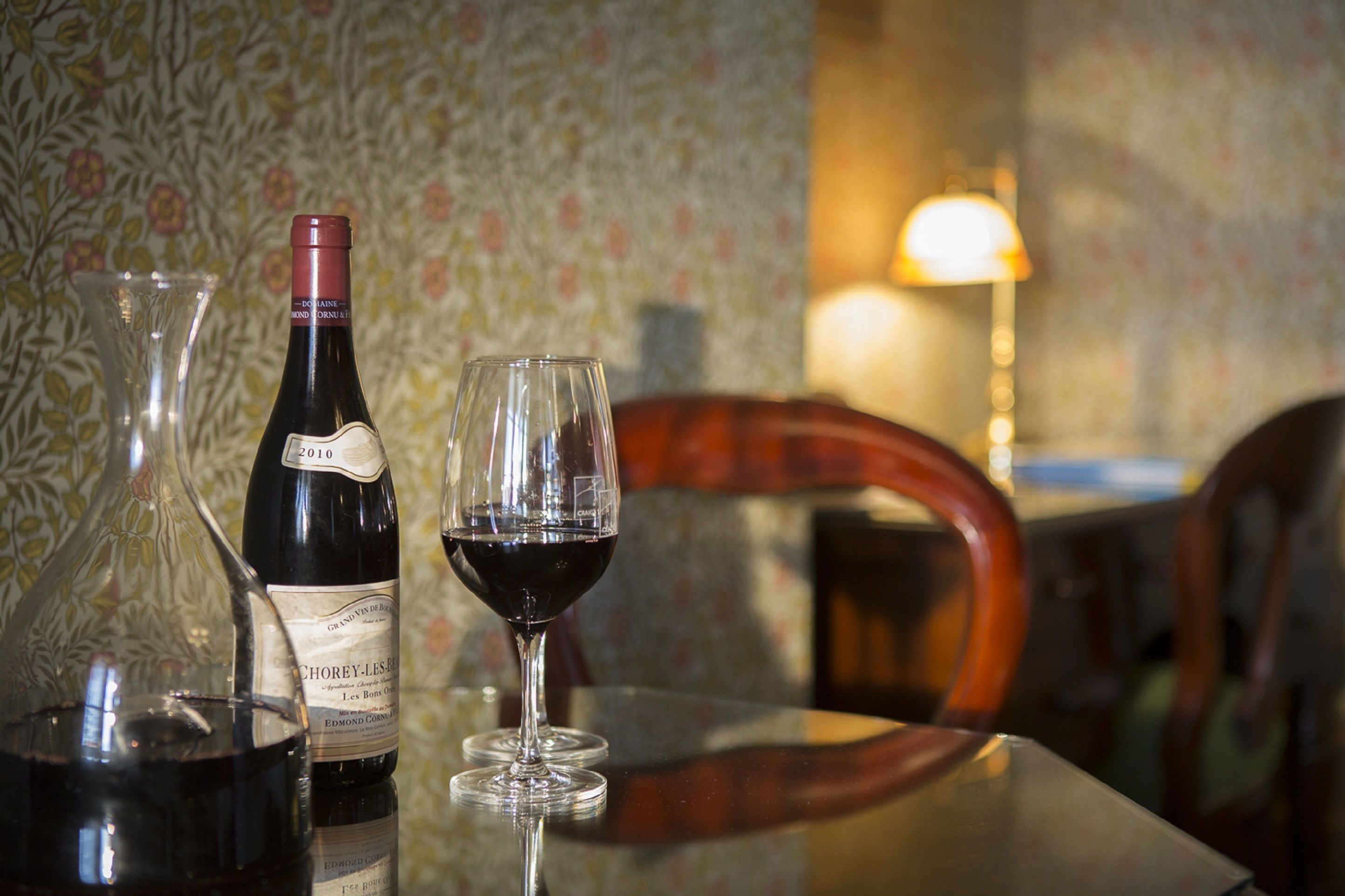 A bottle of wine and a glass on a table with a floral wallpaper background and a lamp.