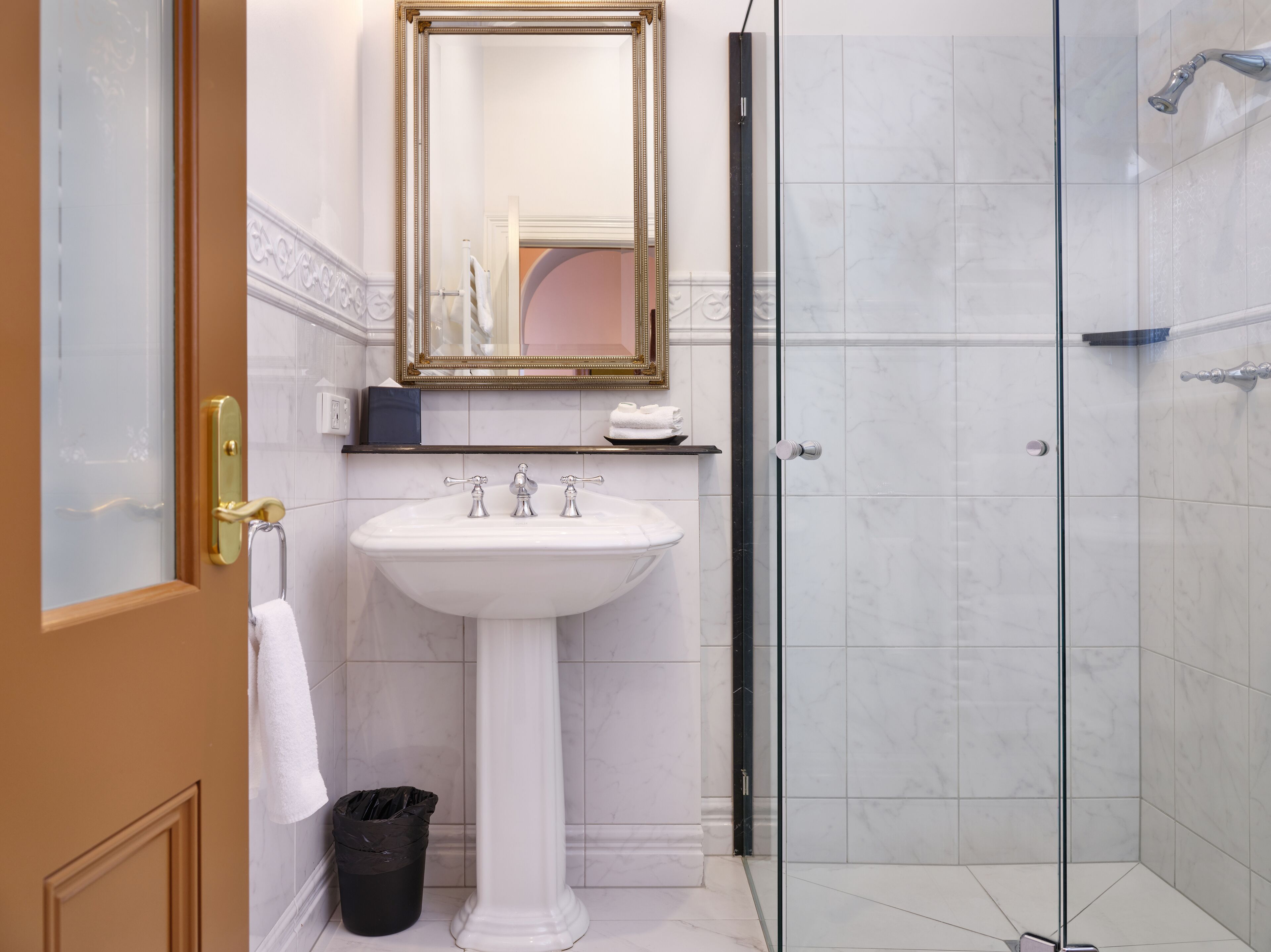 Un baño con lavamanos pedestal blanco, cabina de ducha de vidrio y espejo con marco dorado.