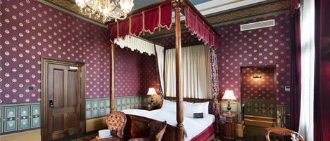 A four-poster canopy bed with a wooden frame and red canopy, a sitting chair, a table with a vase, and a chandelier.